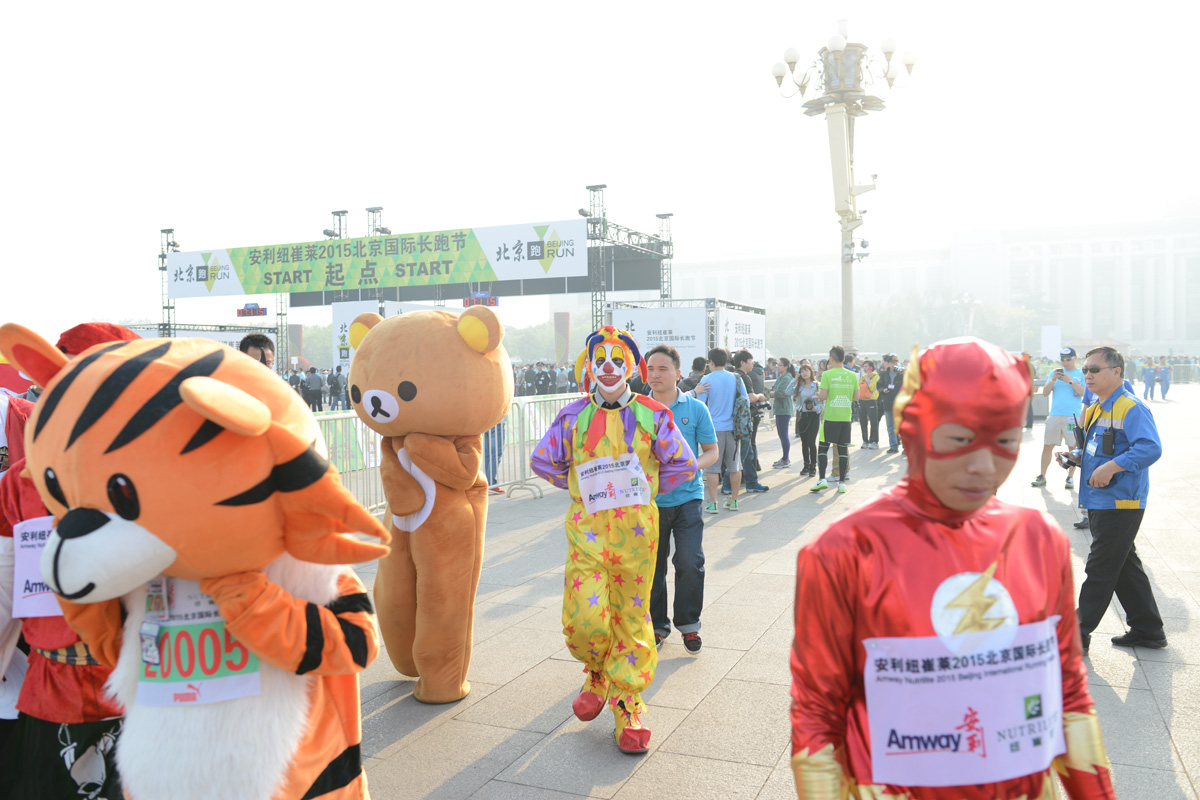 画面为安利纽崔莱2015北京国际长跑节起点区域，背景拱门标注“START 起点”字样。多名参赛者身着老虎、轻松熊人偶服、小丑装、闪电侠造型等趣味装扮候场，号码布印有Amway、纽崔莱标识，周边群众云集，现场氛围活力热闹，尽显健康积极的活动调性。