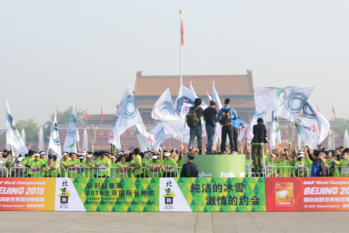 活动背景为天安门广场，围挡标注“安利纽崔莱2015北京国际长跑节”“北京跑”“2015国际田联世界田径锦标赛”等字样。大量身着绿色参赛服的参赛者聚集于此，现场旗帜飞扬，参赛者热情高涨，氛围活力满满，尽显健康向上的运动风貌。