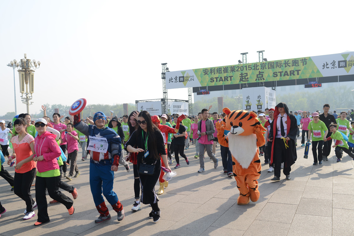 画面为安利纽崔莱2015北京国际长跑节起点场景，大批跑者正从标有“START 起点”的活动拱门出发，队伍中不乏美国队长、老虎人偶等趣味装扮参与者，现场氛围活力热烈，充分传递出健康向上的运动理念。