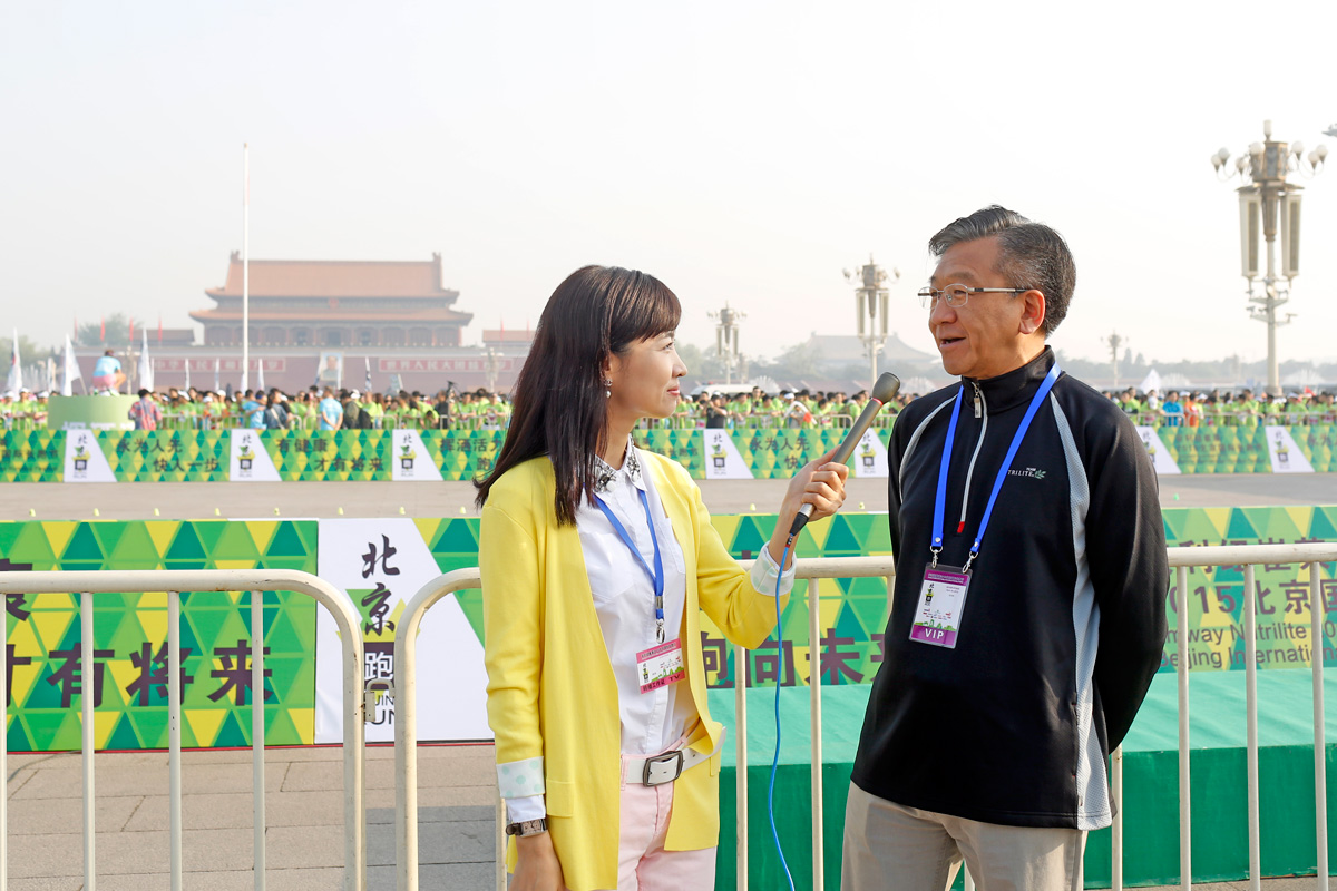 这是2015年安利纽崔莱赞助的北京马拉松活动现场，背景为天安门广场，可见大量参赛人员，围挡印有“有健康才有将来”“北京跑”等标识。女记者正手持话筒采访佩戴VIP证件的嘉宾，现场氛围活力昂扬，展现安利倡导健康生活的品牌理念。