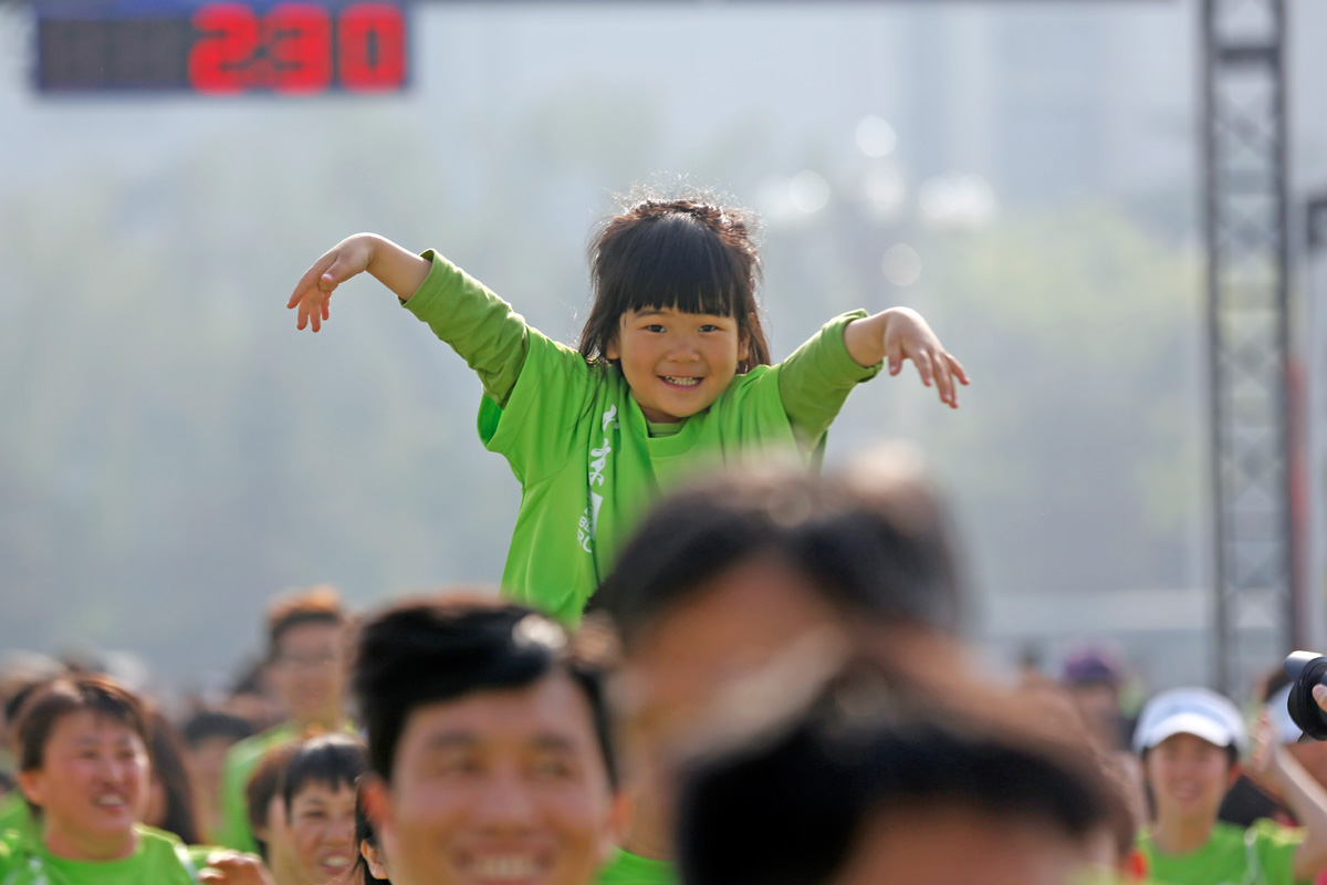 安利健康跑活动现场，参与者身着统一绿色活动服，一名小女孩被托举在人群中，张开手臂笑容灿烂，背景计时牌显示数字“230”，全场参与者情绪饱满，氛围欢快热烈，尽显安利健康向上的品牌活力。