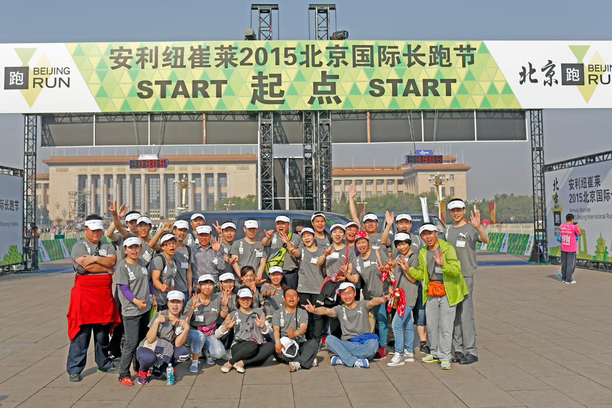 画面位于安利纽崔莱2015北京国际长跑节起点拱门处，数十名身着统一参赛服饰的人员聚集在此合影，大家笑容灿烂，纷纷比出活力手势，现场氛围热烈昂扬，充分展现出活动健康向上的风貌，传递安利倡导的健康生活理念。