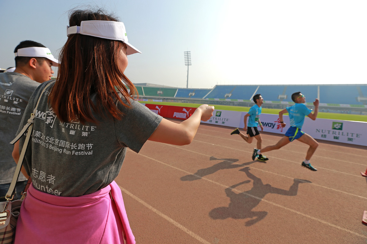 这是2015纽崔莱北京国际长跑节活动现场，身着印有活动标识的安利志愿者在跑道旁为选手加油鼓劲，参赛选手在跑道上奋力向前奔跑，场边围挡标注有纽崔莱、安利品牌标识，现场氛围昂扬热烈，尽显健康向上的运动活力。