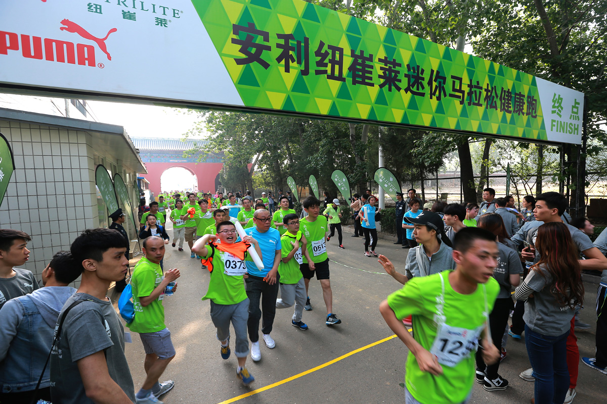 画面呈现安利纽崔莱迷你马拉松健康跑终点场景，拱门印有“安利纽崔莱迷你马拉松健康跑 终点FINISH”及纽崔莱、PUMA标识，身着统一绿色参赛服的参赛者正陆续冲线，周边工作人员、观赛人员驻足，现场氛围热烈活力，尽显安利倡导的健康运动理念。