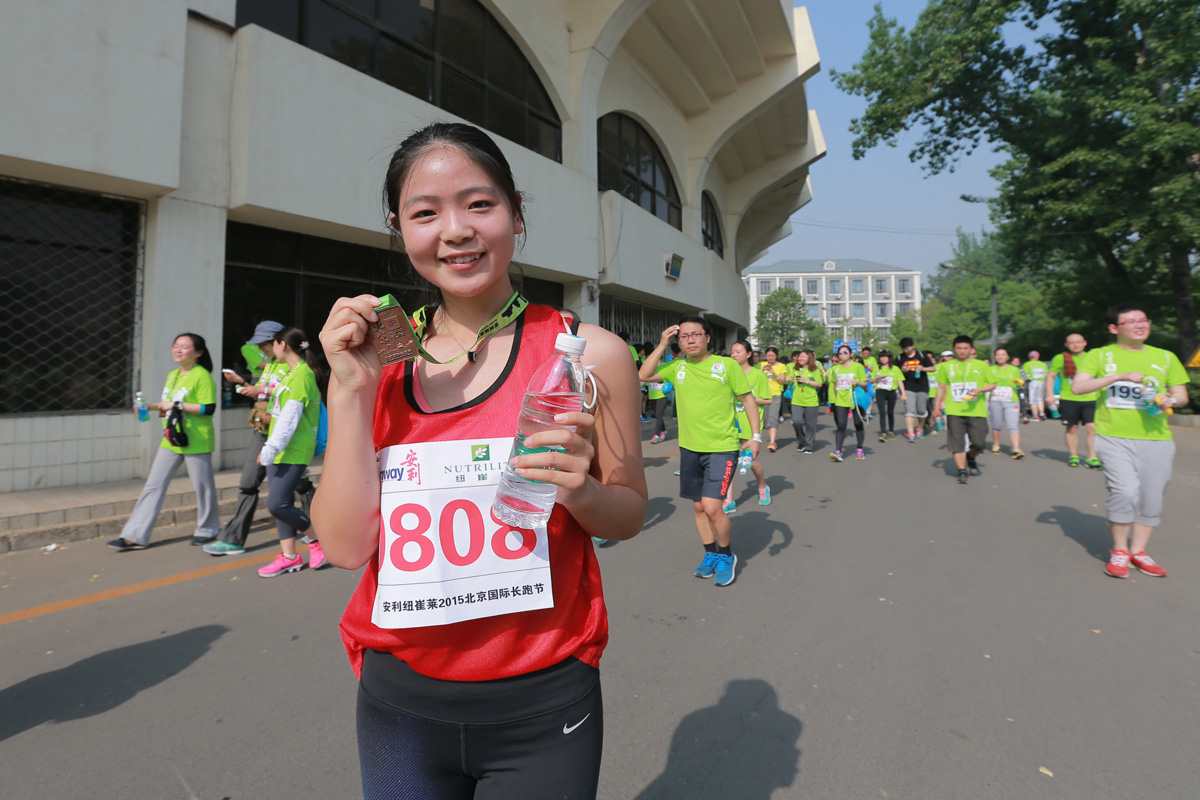 本次为安利纽崔莱2015北京国际长跑节活动现场，佩戴0808参赛号码布的女选手完赛后手持奖牌与矿泉水，笑容明亮，身后大批身着统一绿色参赛服的跑者同行，现场氛围活力昂扬，传递出健康向上的运动理念。