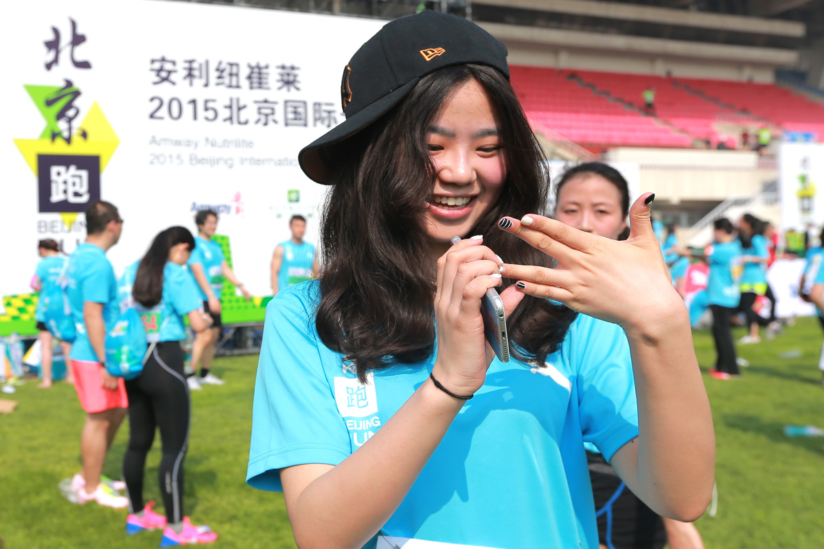 本次为2015安利纽崔莱北京国际跑活动现场，身着统一蓝色活动服的女参与者笑容明媚，正开心摆弄手机，背景是活动主题展板，草坪上还有不少同场参与活动的人员，现场氛围轻松热烈，尽显健康活力的运动氛围，契合安利健康向上的品牌调性。