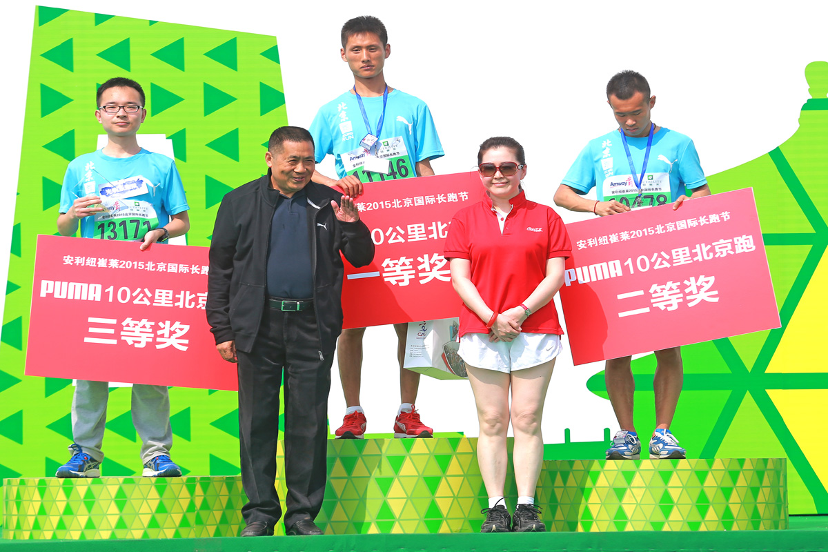 本场景为安利纽崔莱2015北京国际长跑节PUMA10公里北京跑颁奖环节，三位获奖选手站在领奖台上，分别手持对应一、二、三等奖的红色标识牌，两位颁奖嘉宾立于台前，现场氛围昂扬热烈，充分传递出健康向上的运动活力，契合安利的健康品牌调性。