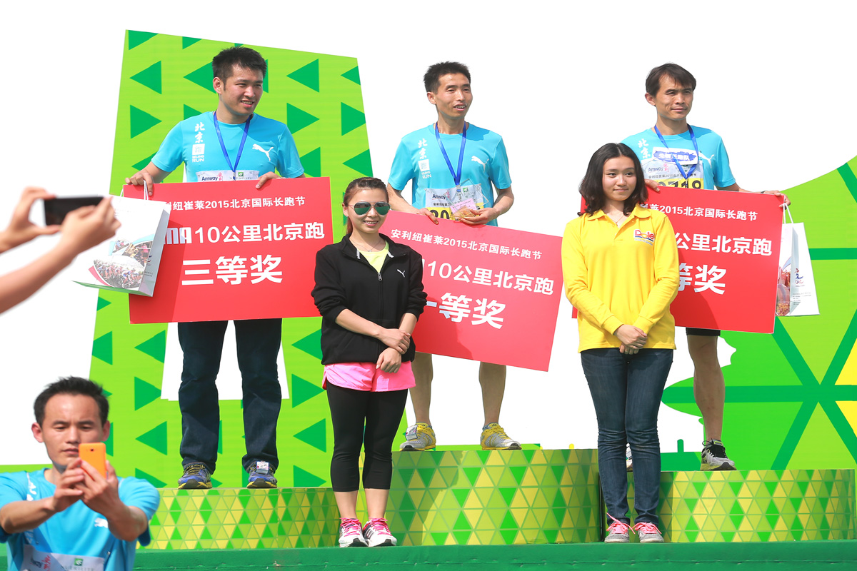 画面为安利纽崔莱2015北京国际长跑节10公里跑颁奖现场，获奖选手站在颁奖台上，分别手持标注一等奖、二等奖、三等奖的红色展示牌，现场有人员拍照记录，氛围热烈昂扬，彰显运动赛事的活力与正向价值。