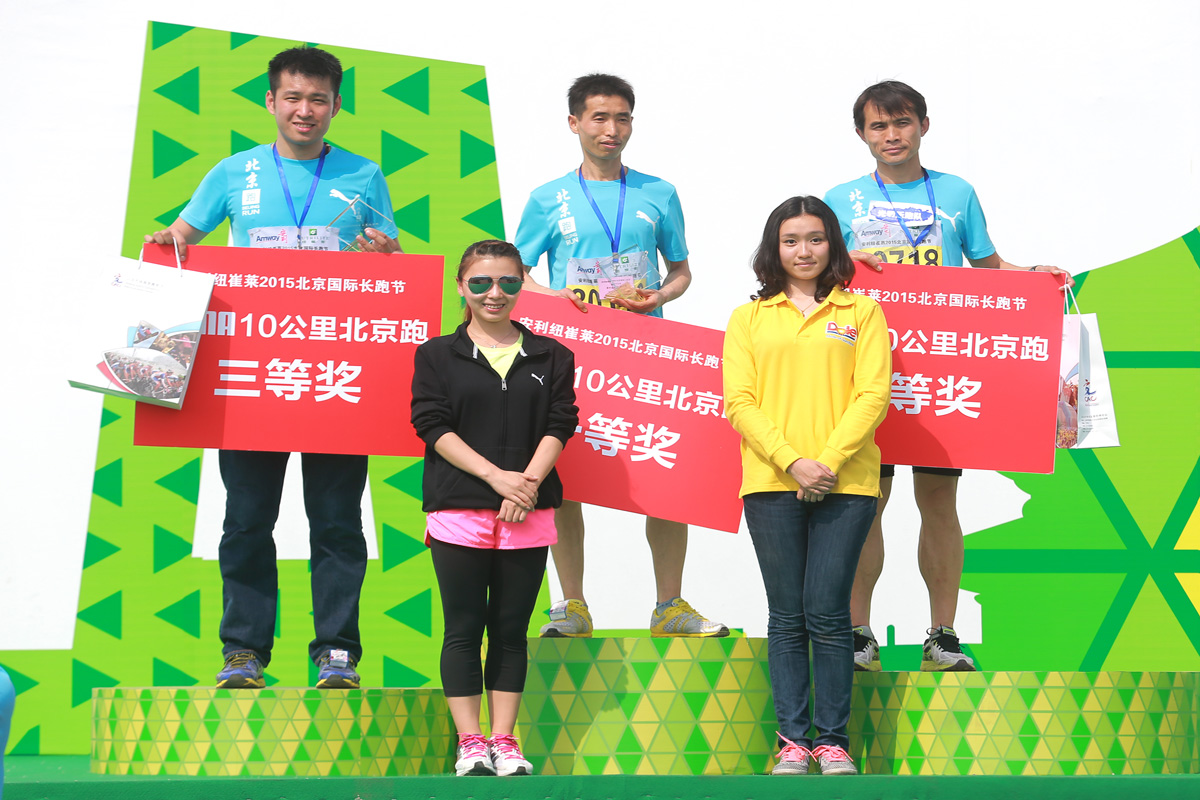 画面为安利纽崔莱2015北京国际长跑节10公里跑颁奖现场，三名获奖选手站在黄绿风格领奖台上，分别手持对应等级的奖项标识、奖牌与奖品，两名颁奖嘉宾站立于台前，现场氛围积极昂扬，传递出健康向上的运动理念与品牌活力。