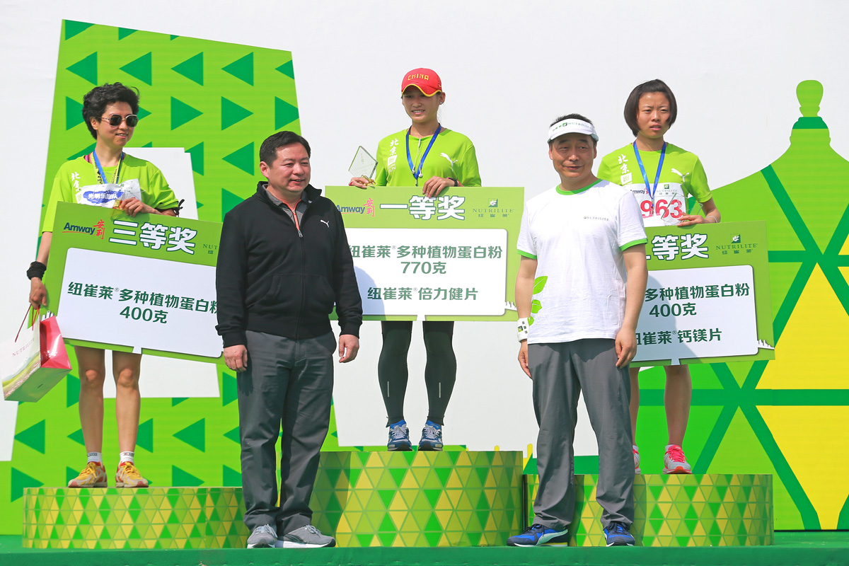画面为安利纽崔莱健康跑颁奖场景，两位嘉宾与三位获奖女选手站在绿黄主题颁奖台上，三位选手分别持有对应一二三等奖的奖牌，奖品包含纽崔莱多种植物蛋白粉、倍力健片、钙镁片等产品，现场氛围积极明快，传递健康向上的品牌理念。
