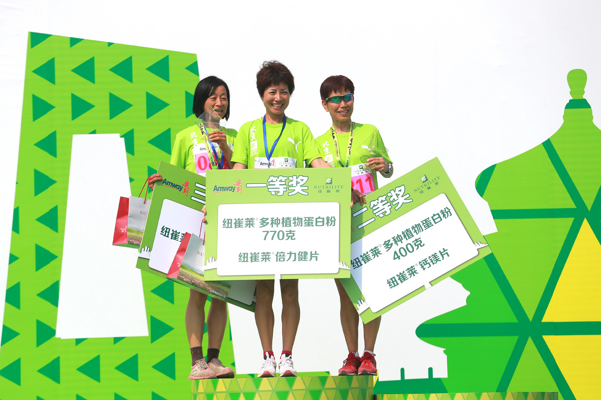 图为安利纽崔莱跑步赛事领奖现场，三位身着绿色参赛服的女性获奖选手站在领奖台上，分别持有对应一二三等奖的奖项牌与奖杯，一等奖奖品为纽崔莱770克多种植物蛋白粉、倍力健片，二等奖含400克纽崔莱蛋白粉、钙镁片，现场氛围昂扬喜悦，尽显健康活力。