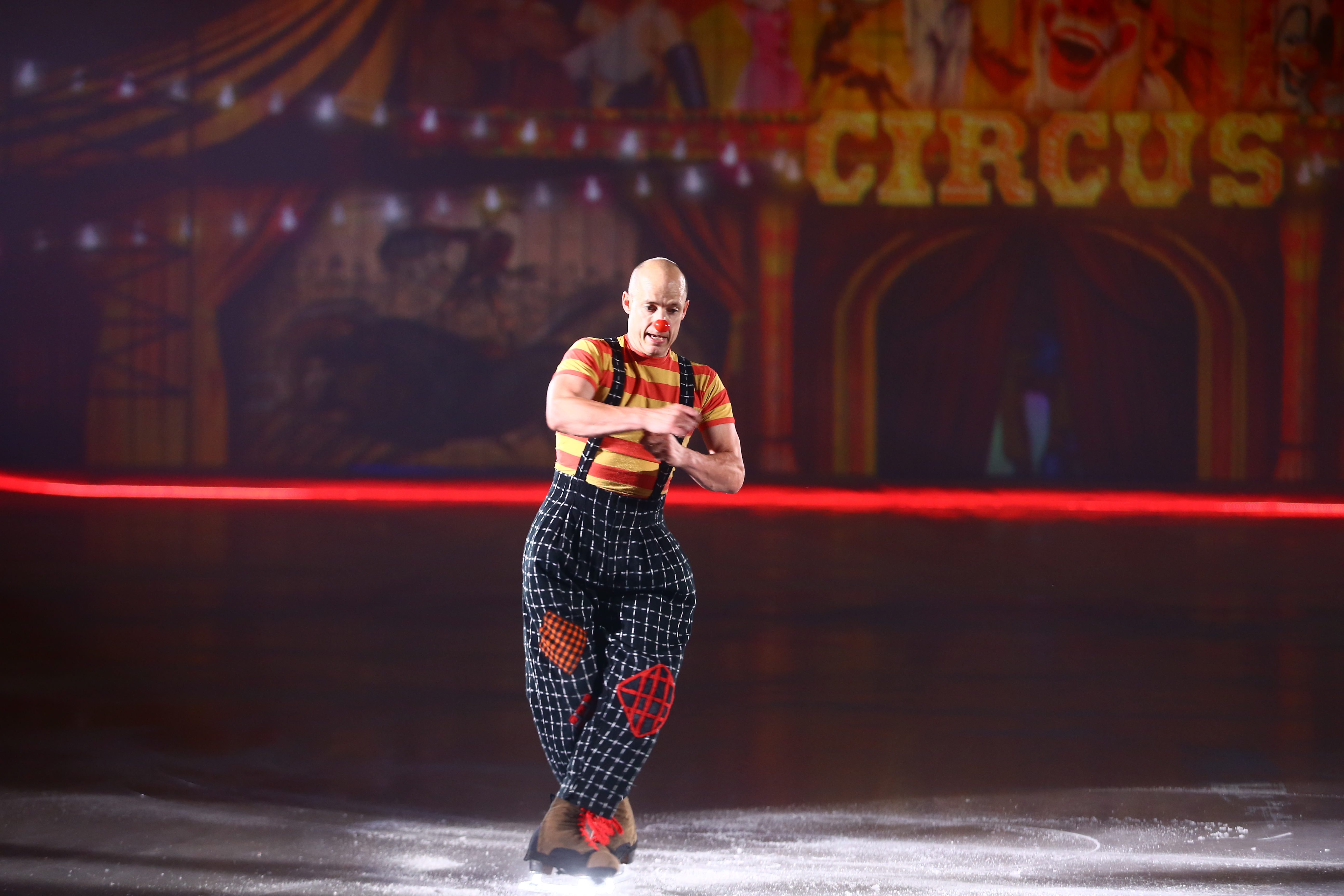 本次安利品牌活动现场，背景带有核心字样“CIRCUS”的马戏主题布景下，佩戴红鼻头、身着红黄条纹上衣与格纹背带裤的小丑演员正在冰面进行趣味滑冰演出，诙谐生动的表演营造出轻松欢快的氛围，为到场伙伴带来趣味十足的观赏体验。
