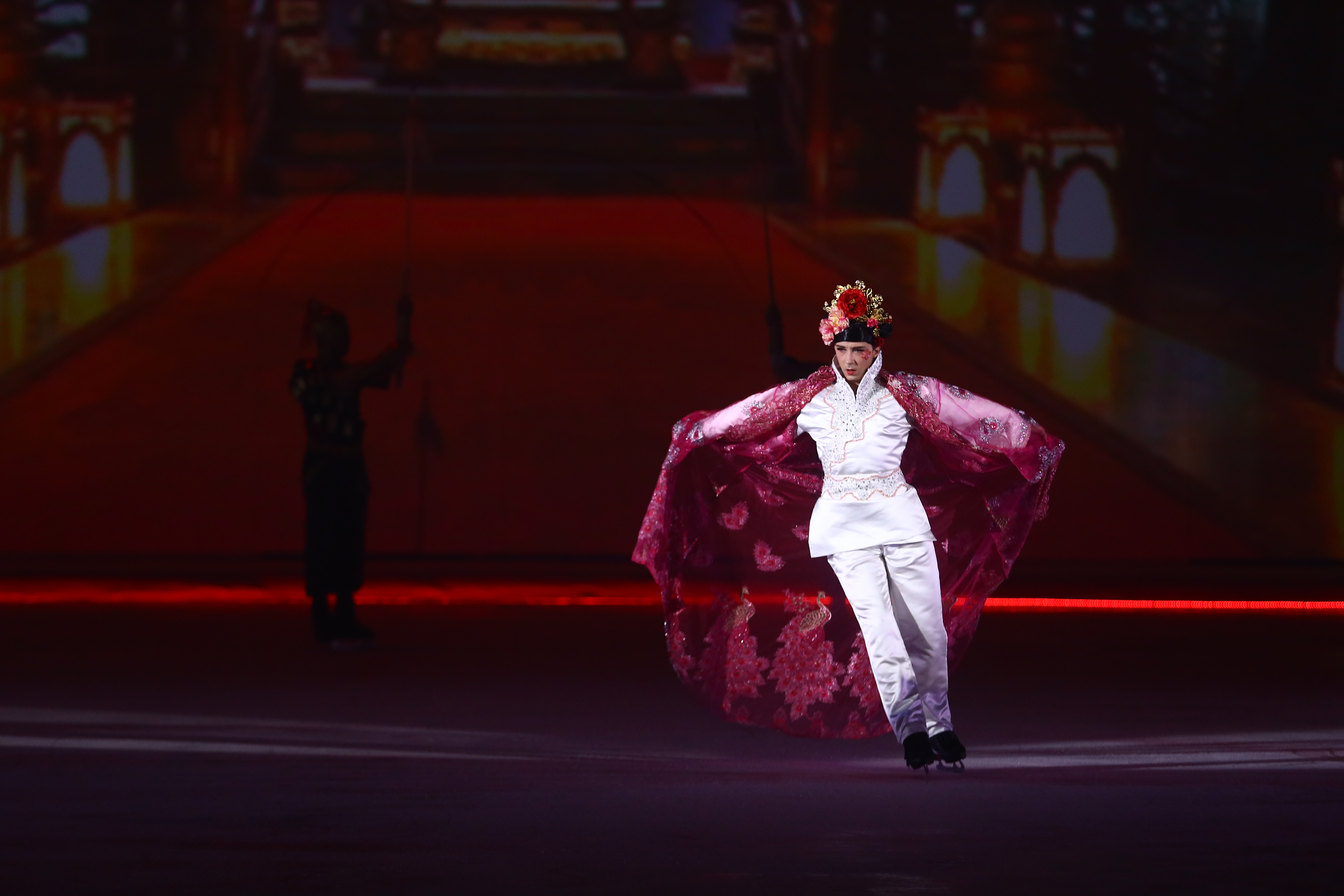 这是安利品牌活动现场的冰上演出场景，表演者头戴华丽珠花头冠，身着白色国风演出服搭配绣有精美纹样的玫红色亮面披风，在冰面舒展滑行，背景搭配古雅的舞台投影，现场氛围绚烂震撼，尽显传统美学与创意演出的融合魅力。