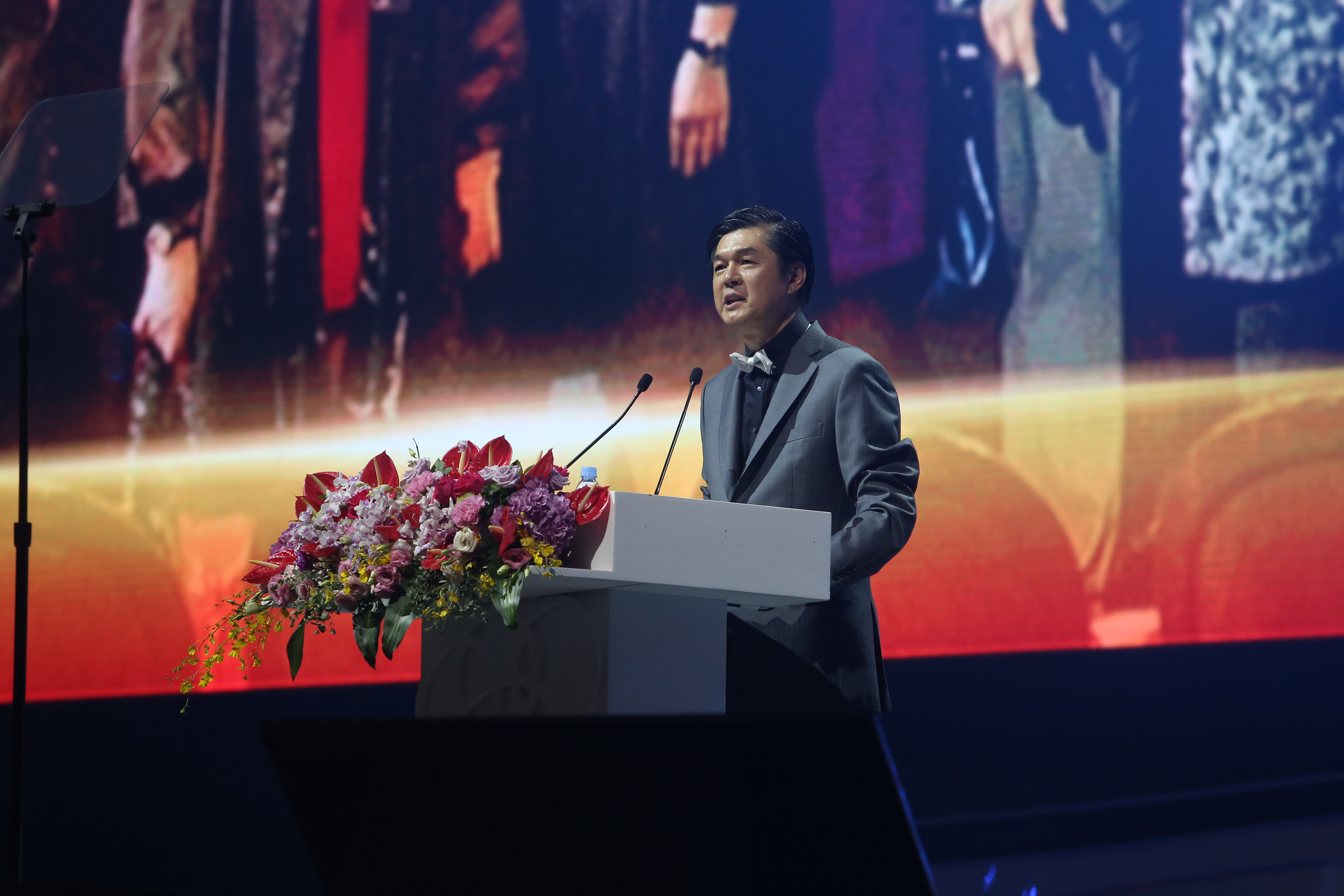 活动现场，身着灰色西装、佩戴白色领结的发言嘉宾站在装饰有多色鲜花的白色讲台后致辞，讲台配有双收音麦，背景是炫彩光影大屏，整体氛围庄重正式，尽显安利品牌活动的专业规格与昂扬风貌。