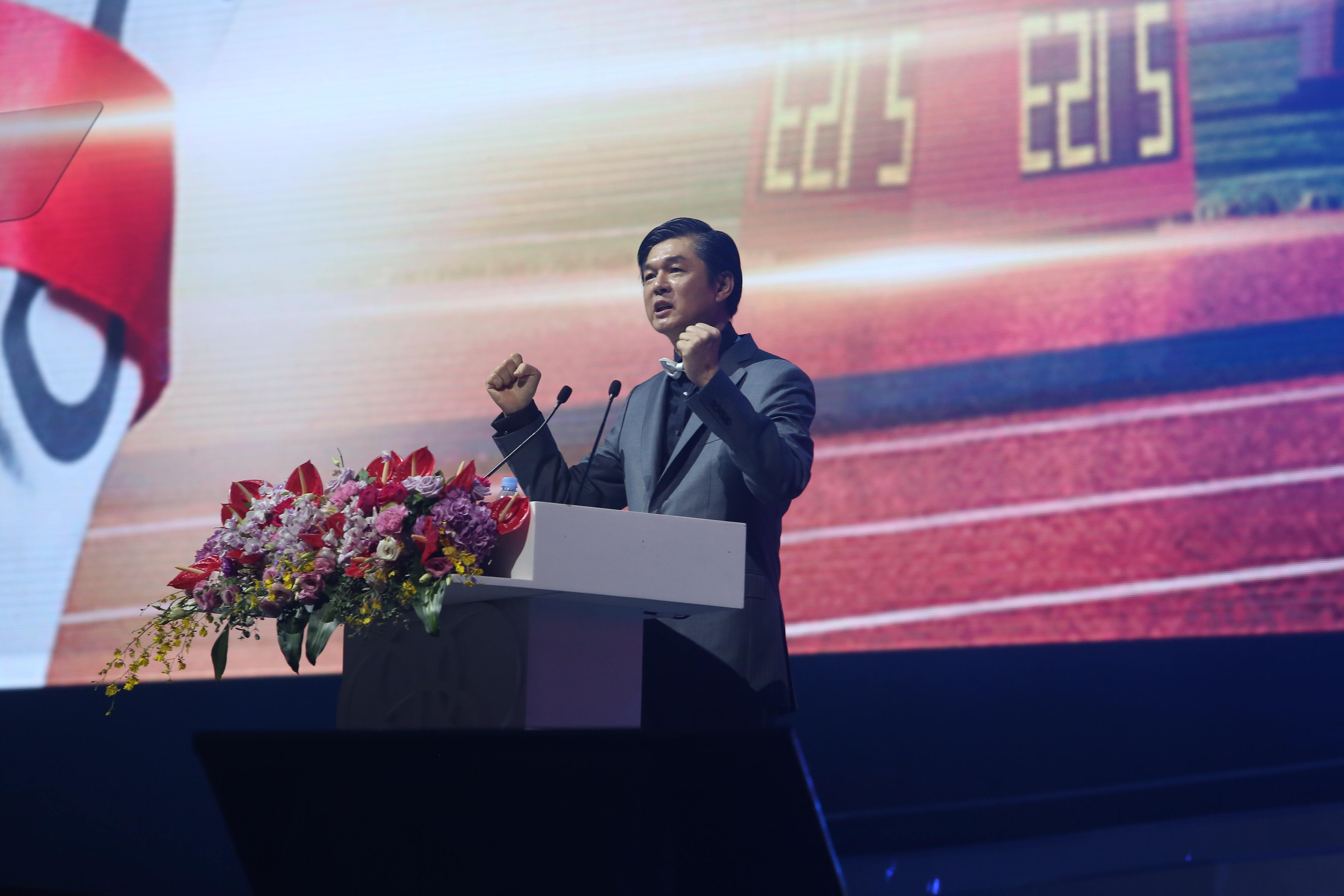 安利品牌活动现场，身着正装的高管站在装饰有精美花艺的演讲台后，握拳激昂发表致辞，背景大屏显示“E215”字样，现场氛围昂扬振奋，传递出锐意进取的发展信心，契合安利积极向上的品牌调性。