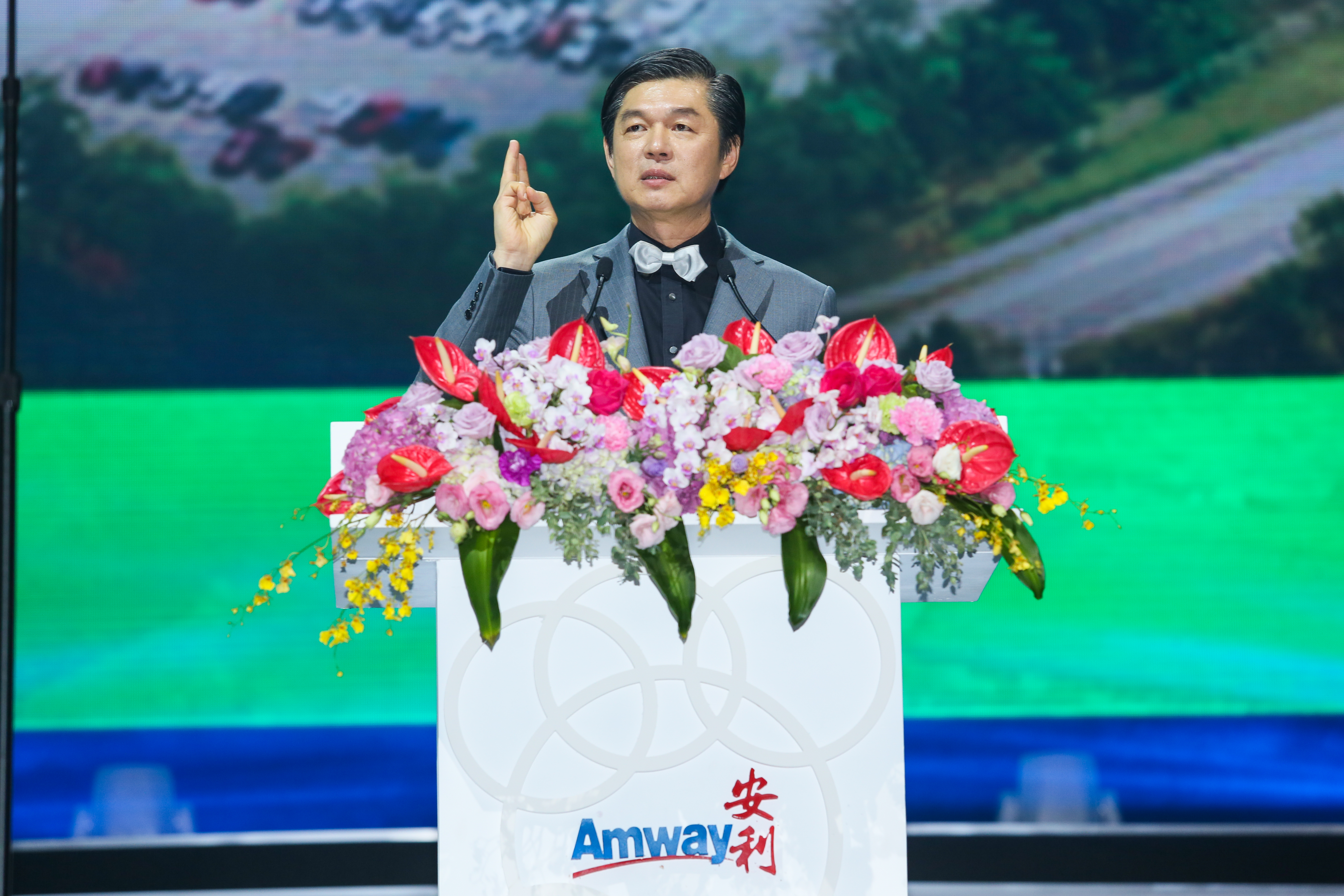 印有“Amway 安利”标识的演讲台装点着多彩鲜切花，身着灰色西装搭配领结的男士站在台后抬手发言，神情从容笃定，背景为活动大屏，现场氛围正式积极，尽显安利品牌活动的专业风貌。