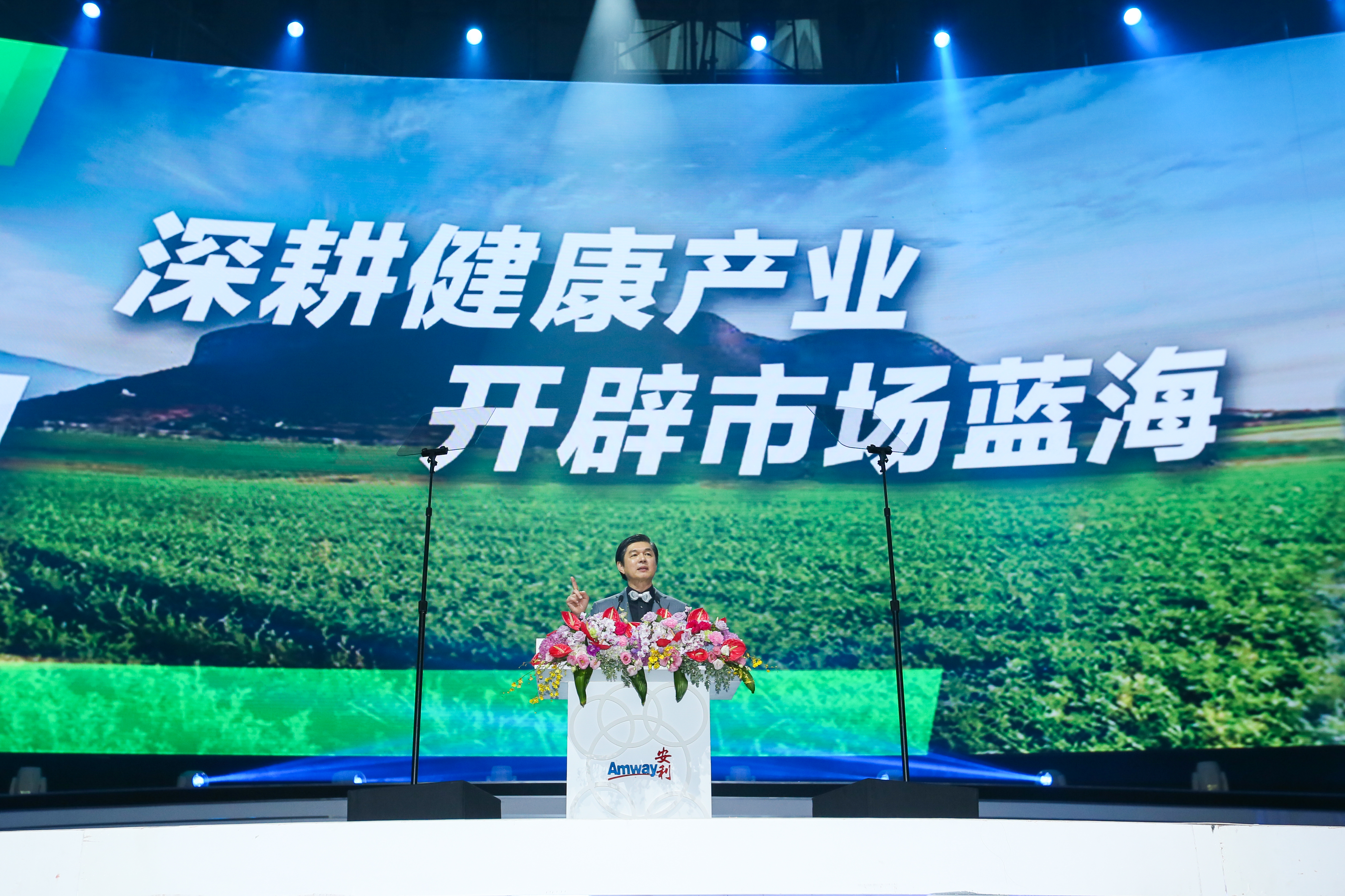 本次安利活动现场，背景大屏醒目展示“深耕健康产业 开辟市场蓝海”字样，发言人立于印有安利Amway标识、装饰着鲜切花的发言台后发表讲话，现场灯光专业明亮，氛围庄重昂扬，传递出安利布局大健康赛道的坚定方向。