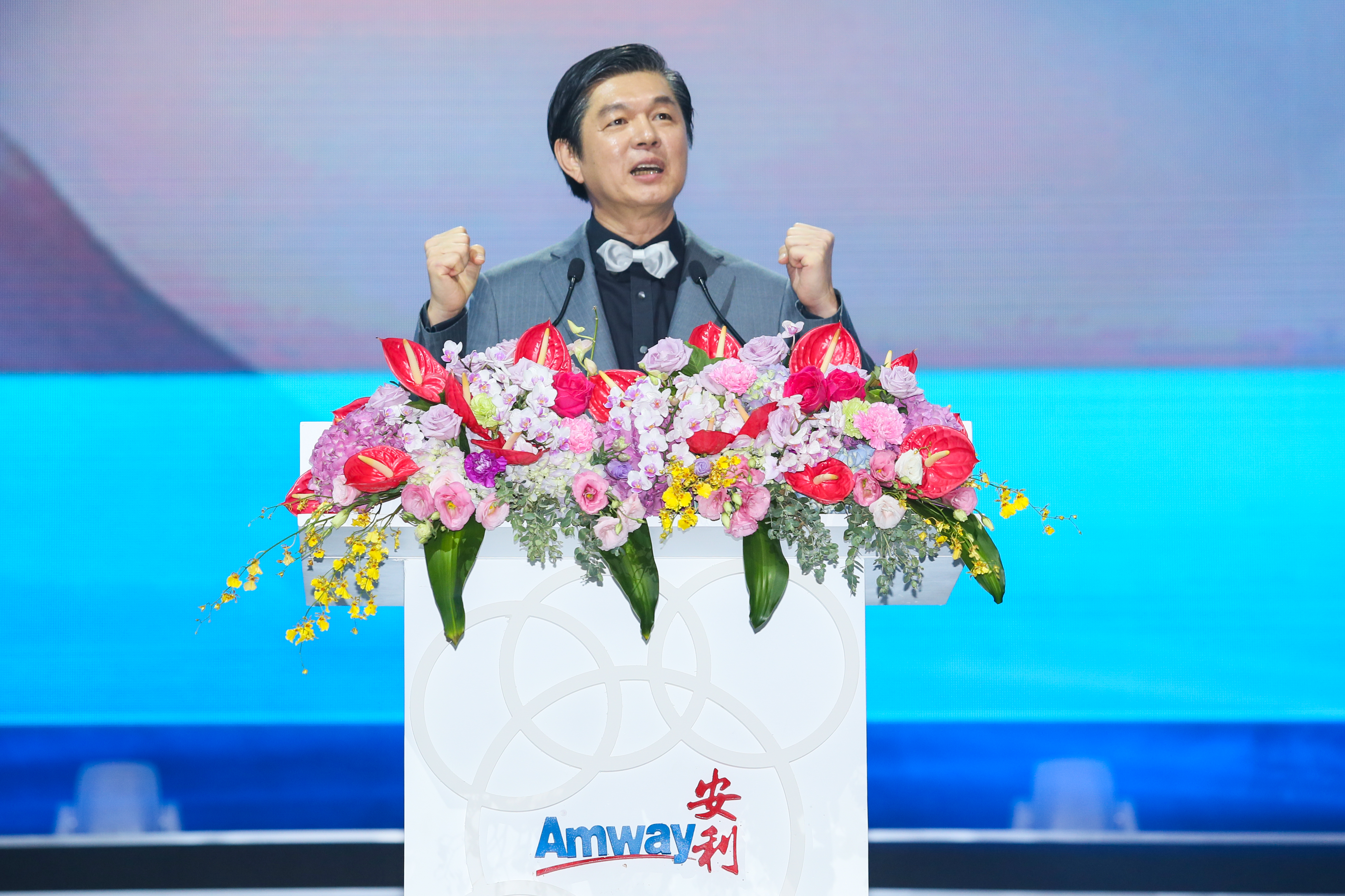 在安利品牌活动现场，印有“Amway 安利”标识的演讲台装饰着色彩丰富的鲜切花，身着灰色西装、佩戴白色领结的嘉宾正于台后发言，他握拳鼓劲的姿态昂扬振奋，现场氛围积极热烈，彰显出安利昂扬向上的品牌风貌。