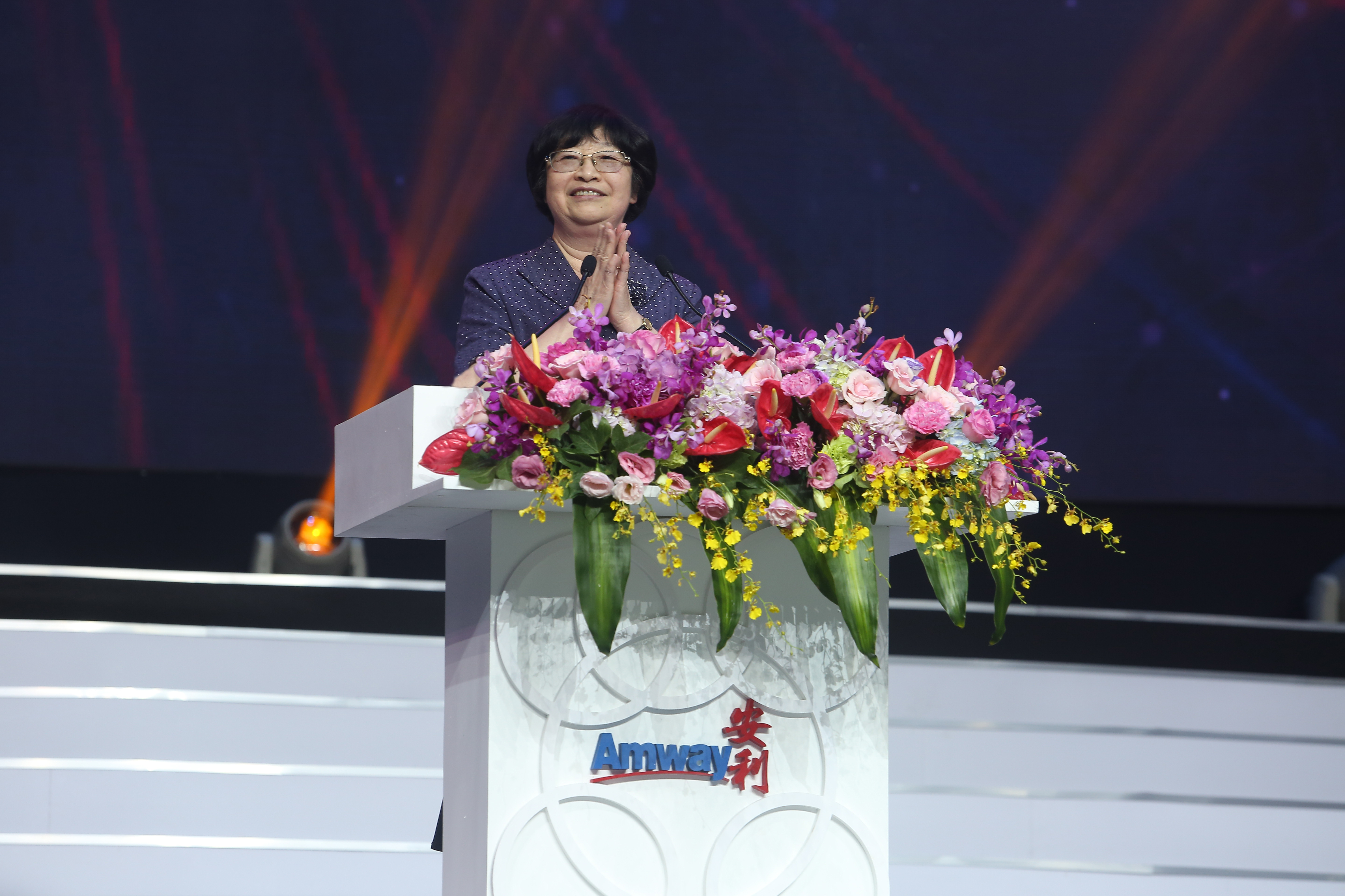 画面为安利（Amway）品牌活动现场，嘉宾立于装饰有缤纷鲜花的品牌讲台后，面带微笑双手合十鼓掌，舞台背景灯光绚丽，整体氛围庄重热烈，尽显活动正式昂扬的基调。
