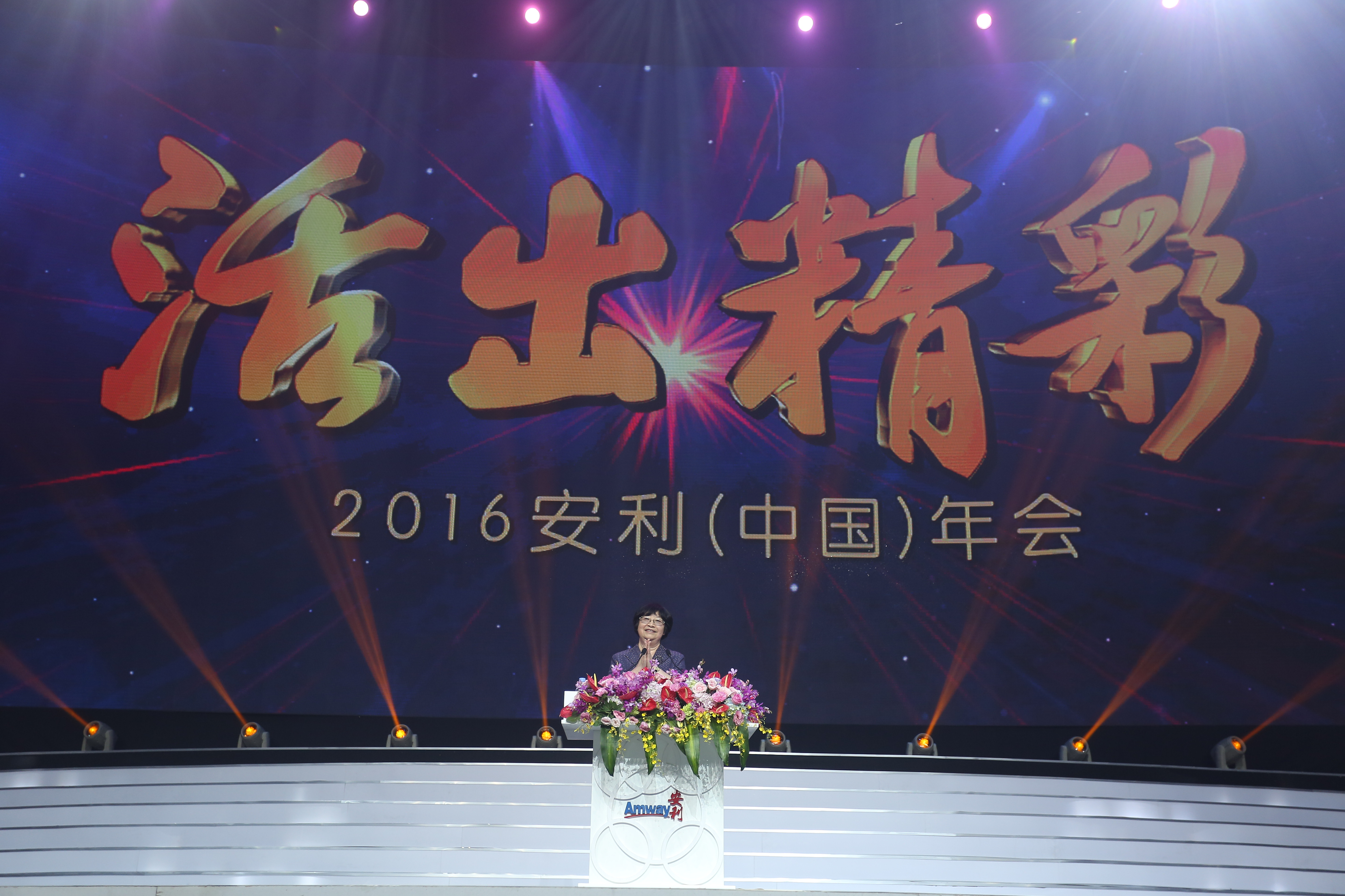 本次场景为2016安利（中国）年会现场，舞台背景大屏醒目呈现“活出精彩”主题与年会全称，现场灯光绚烂氛围隆重。发言人立于装饰鲜花、印有Amway安利标识的讲台后发言，整体氛围热烈正向，彰显安利昂扬的品牌风貌。