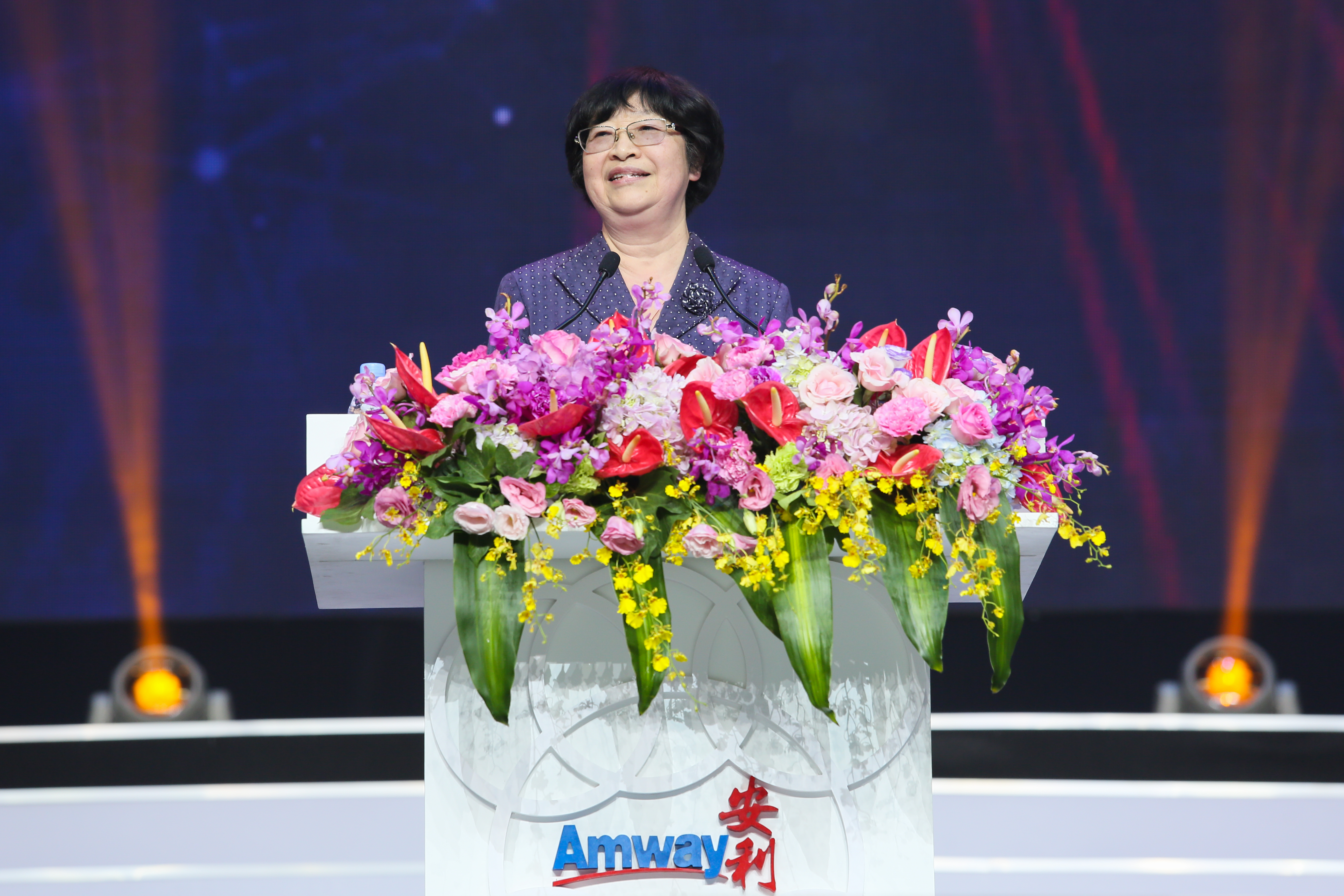 画面中嘉宾站在印有“Amway 安利”标识、装饰着缤纷鲜花的演讲台后发言，舞台背景灯光绚丽柔和，整体氛围正式庄重、积极热烈，尽显安利品牌活动的专业质感与昂扬向上的风貌。
