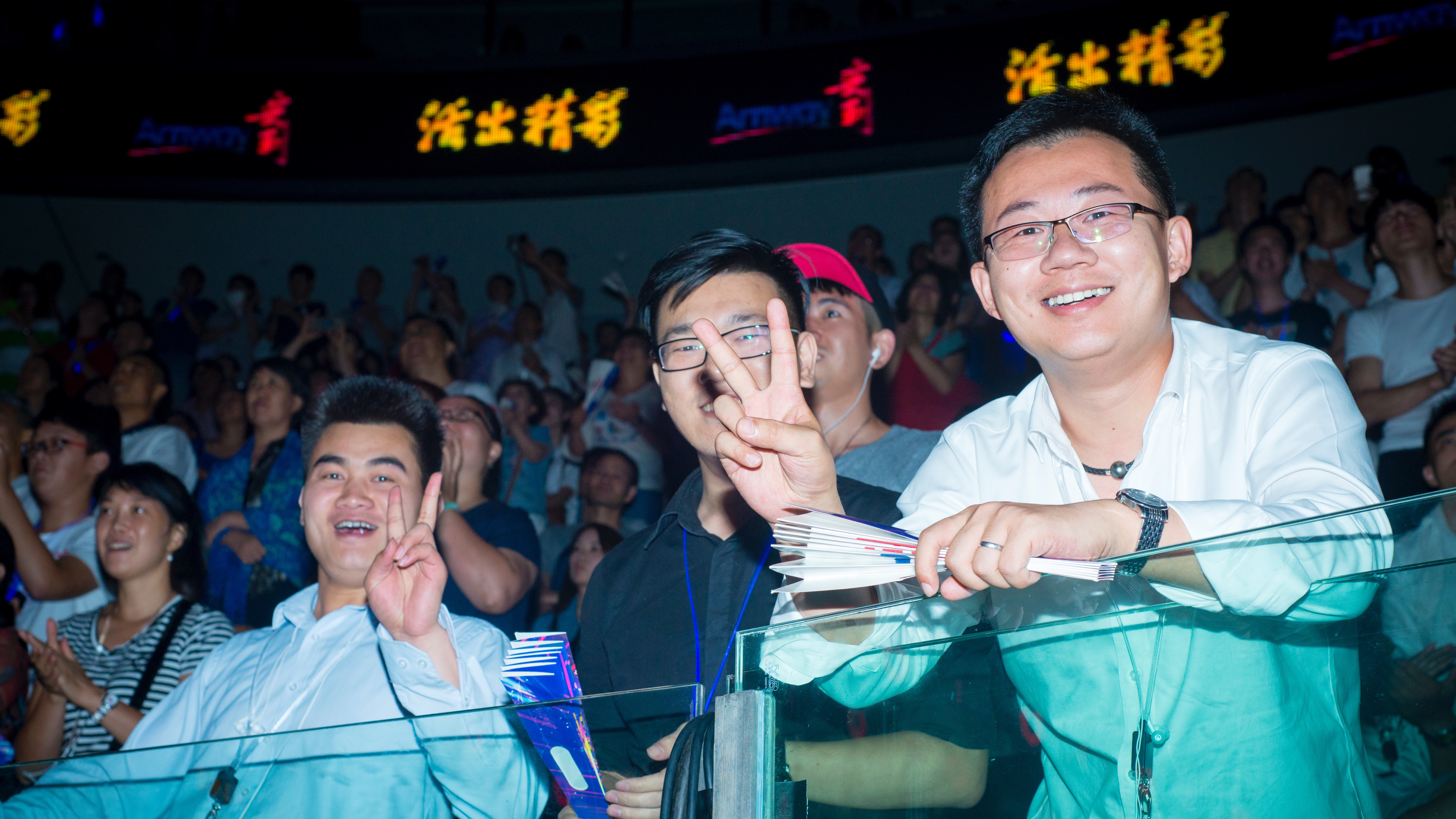 活动现场背景屏显示“活出精彩”与Amway标识，观众席上的伙伴们笑容灿烂，有人比出剪刀手传递喜悦，现场人头攒动、氛围热烈高涨，尽显参与者的昂扬状态，契合安利积极向上的品牌调性。