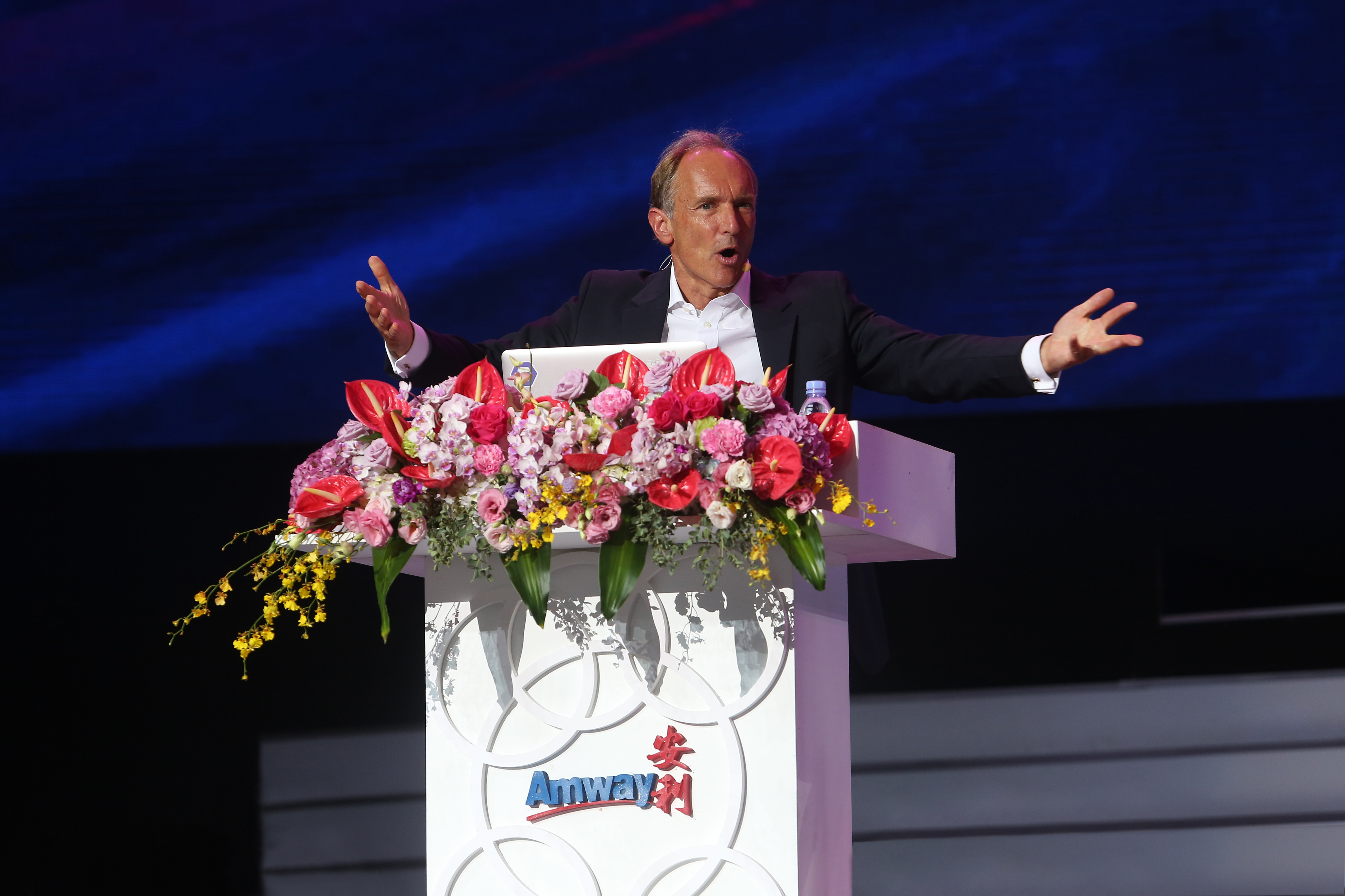 印有“Amway 安利”标识的白色演讲台上装饰着缤纷花束，身着正装的外籍嘉宾站在台后，双臂张开正激情发言，搭配深邃蓝色舞台背景，现场氛围庄重热烈，尽显安利品牌活动的专业昂扬风貌。