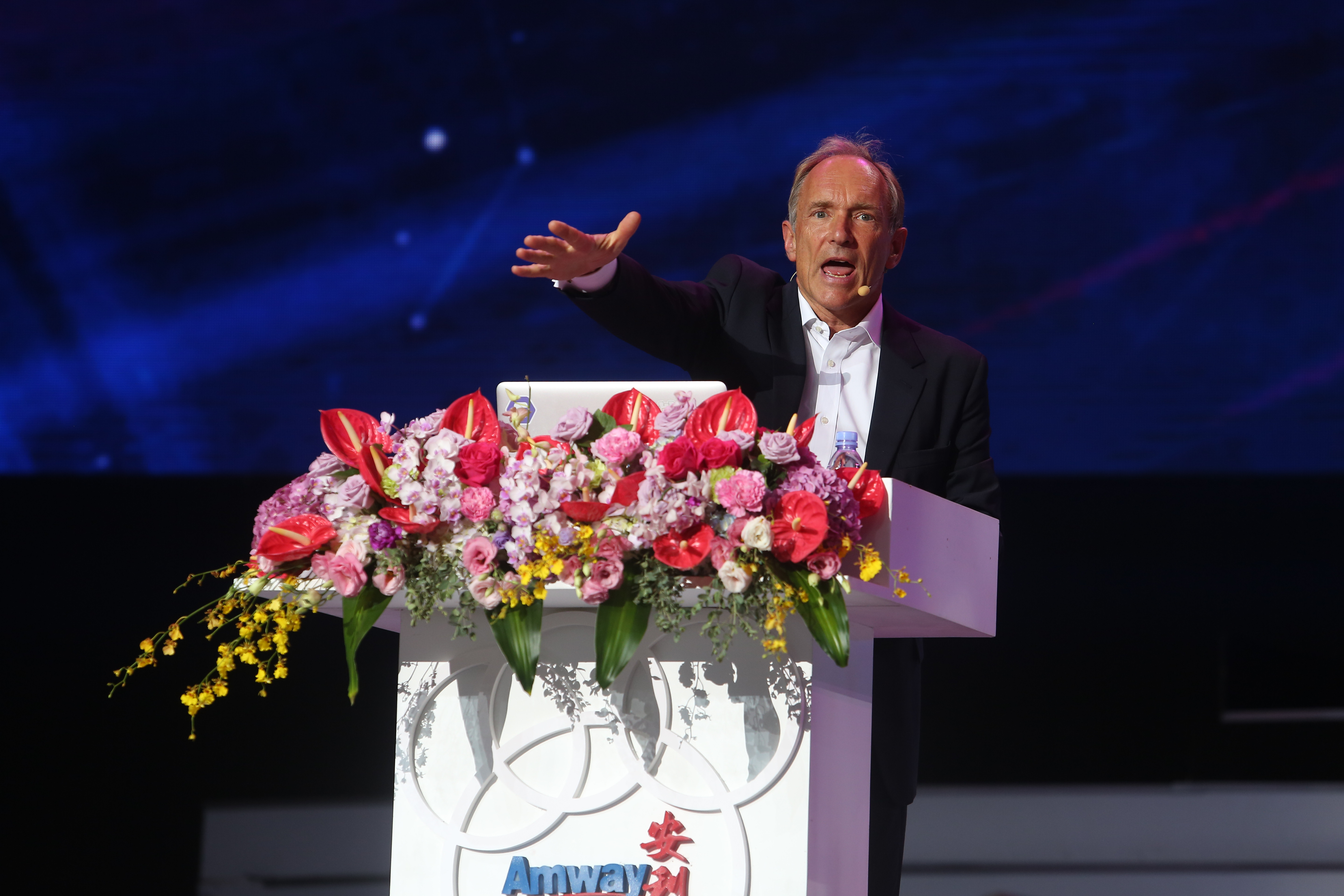 本次安利（Amway）品牌活动现场，外籍嘉宾站在装饰有精美花艺的安利主题演讲台后，正伸展手势激情发言，状态饱满富有感染力，舞台背景为深邃蓝色光影，整体氛围正式热烈，尽显安利高端品牌活动的专业风貌。