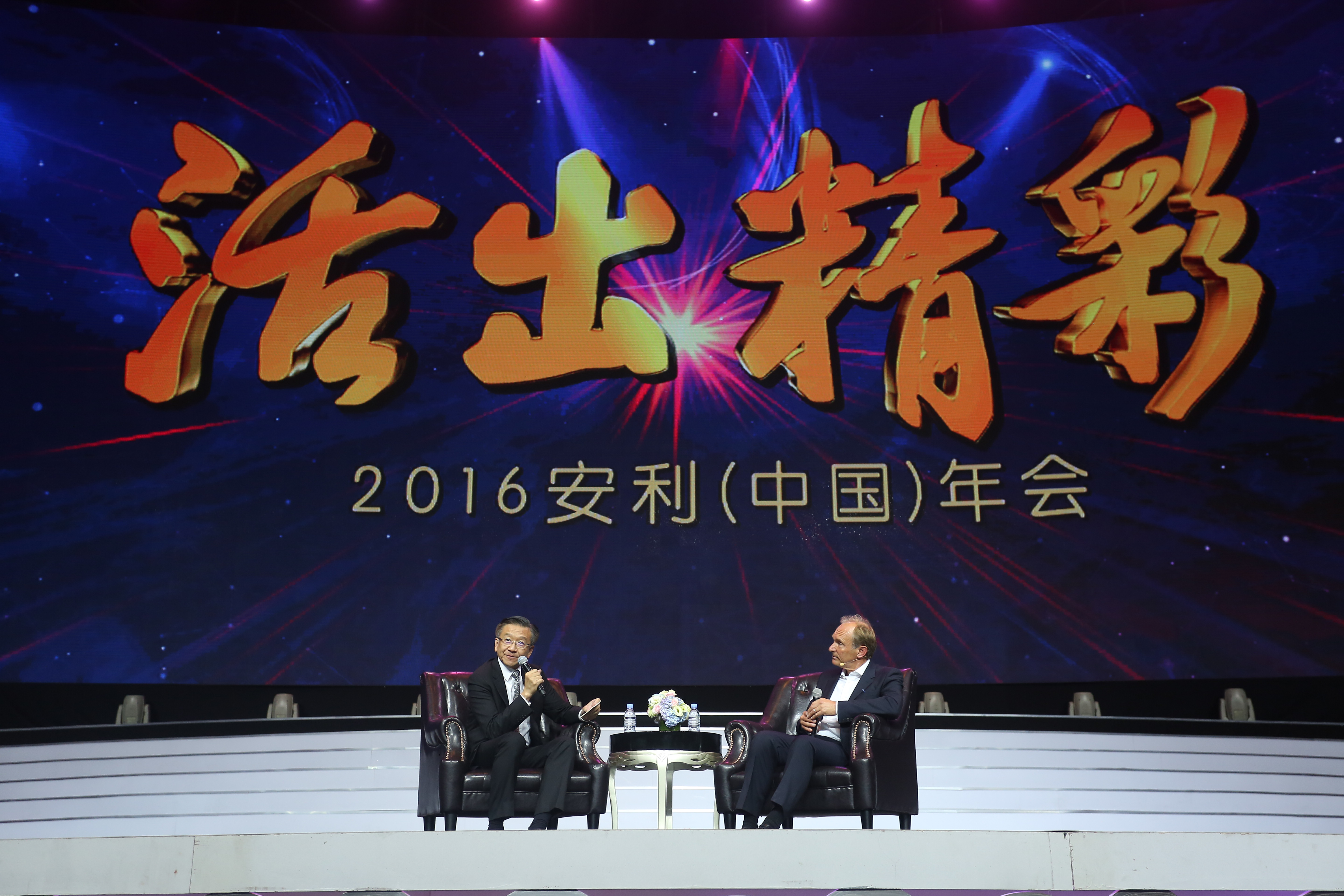 画面为2016安利（中国）年会活动现场，背景大屏醒目展示“活出精彩”主题字样，两位嘉宾落座舞台皮质沙发上开展对话交流，现场灯光绚烂氛围庄重专业，充分展现出安利积极向上、共促发展的品牌风貌。
