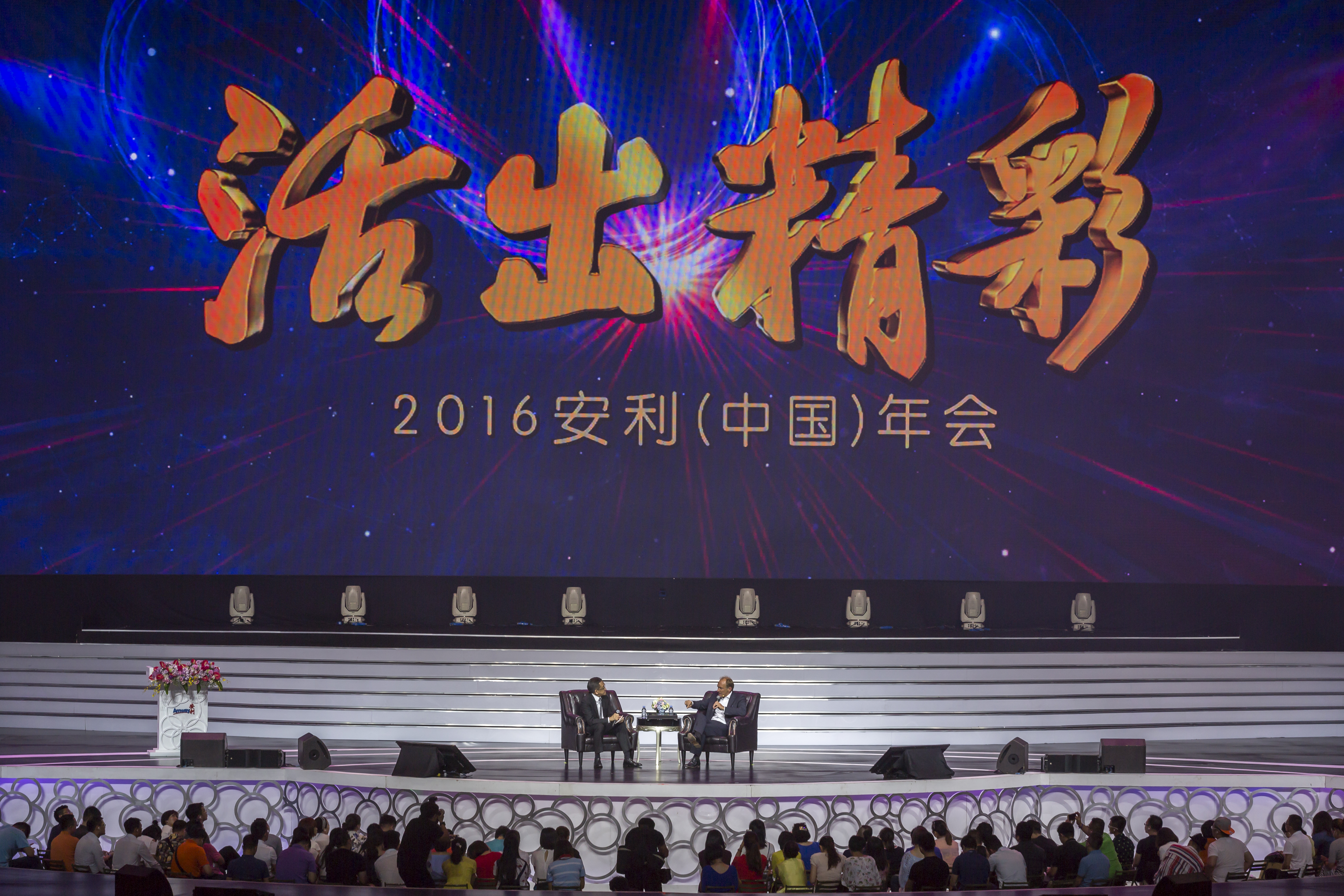 画面为2016安利（中国）年会活动现场，背景大屏醒目展示“活出精彩”主题字样，舞台上两位嘉宾正对坐开展访谈交流，台下坐满参会的安利伙伴，现场氛围热烈庄重，充分彰显出安利积极向上的品牌调性。