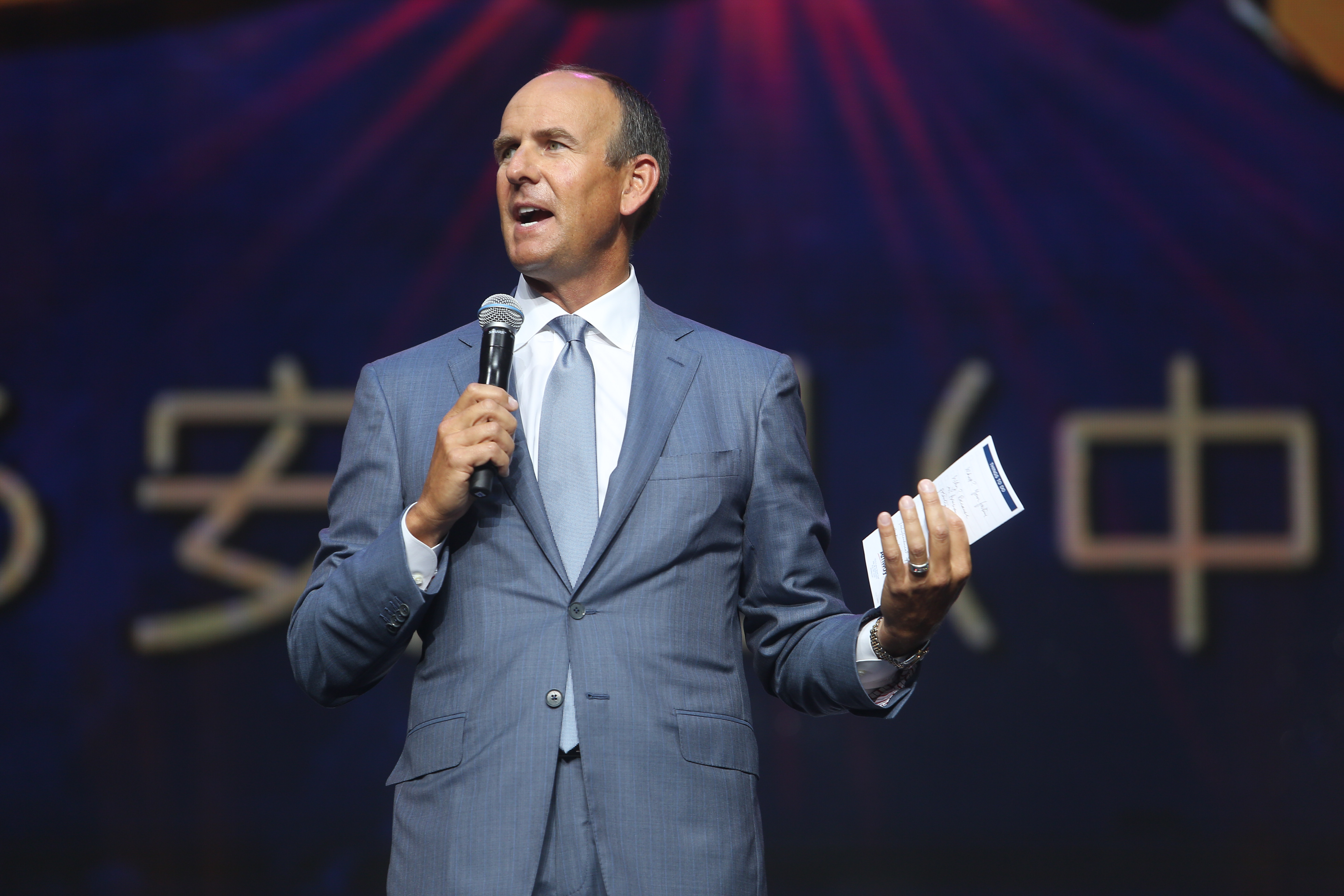 图片为安利中国品牌活动现场，身着浅灰蓝色西装的高管手持麦克风与发言手卡，正站在舞台上发表讲话，背景可见“安”“中”字样标识，现场氛围庄重正式，高管发言状态饱满昂扬，极具正向感召力。
