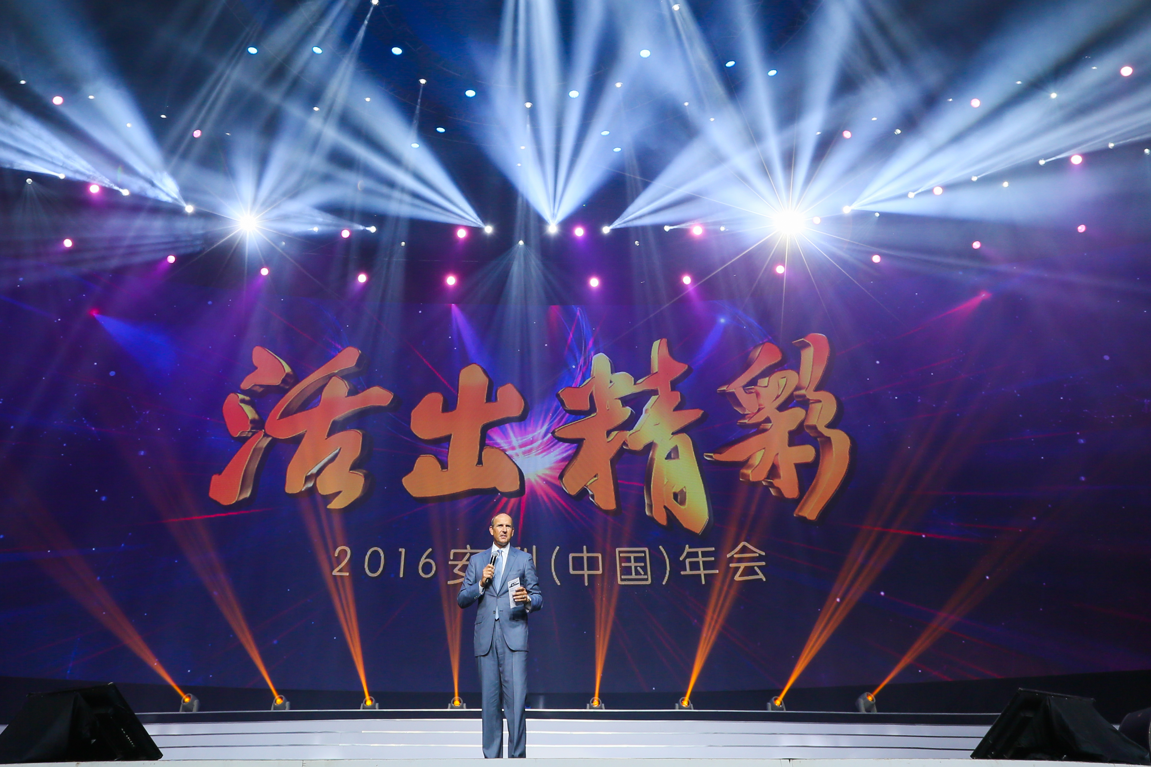 本次是2016安利（中国）年会活动现场，舞台灯光绚烂璀璨，背景大屏醒目展示活动主题“活出精彩”。身着正装的外籍嘉宾手持话筒与手卡站在舞台中央发言，现场氛围隆重热烈，尽显积极向上的品牌面貌。