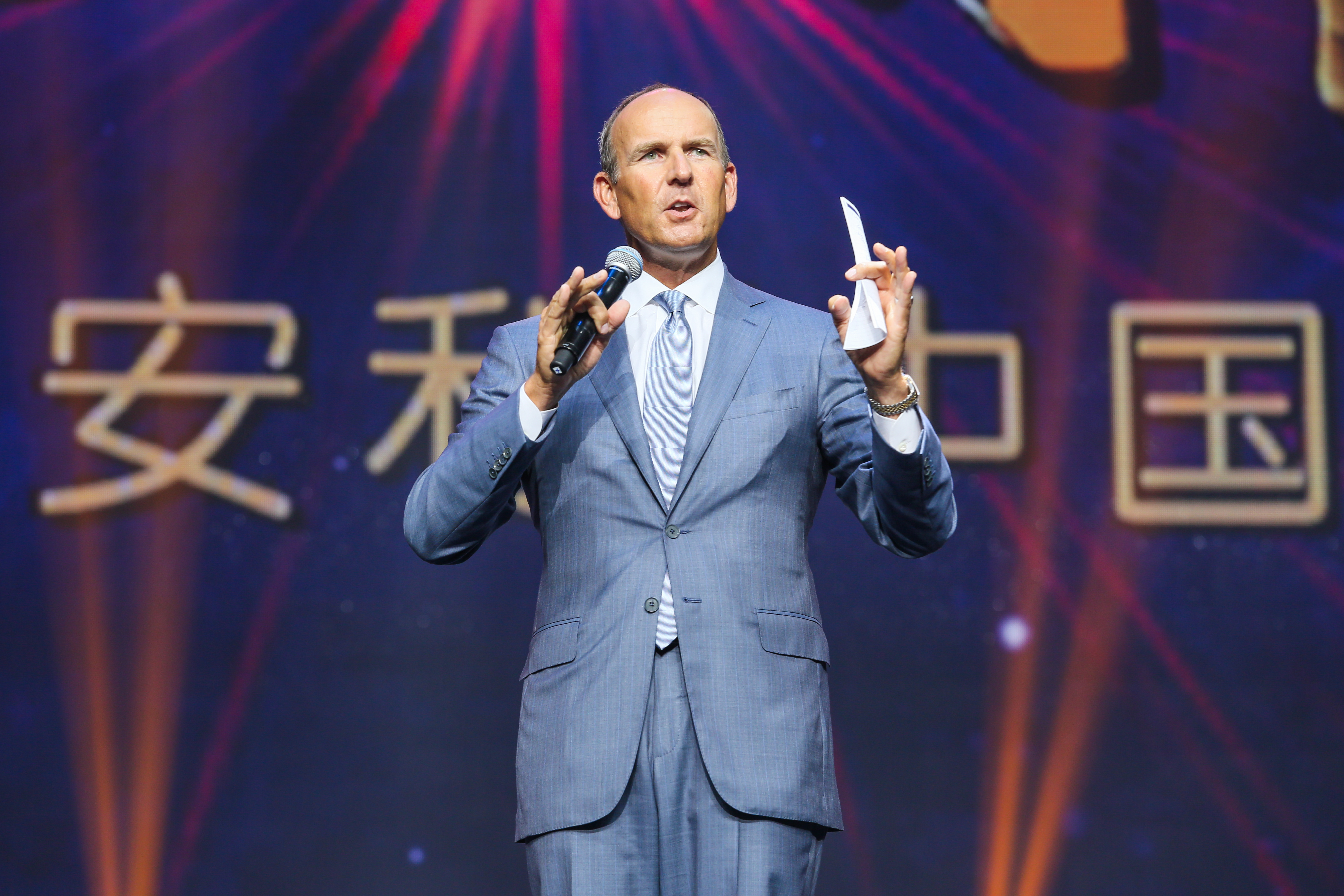 活动现场背景醒目呈现“安利中国”字样，身着灰蓝色西装的外籍高管手持麦克风与手卡，正站在舞台上发言，神情专注、语态从容，搭配绚烂大气的舞台灯光，整体氛围庄重热烈，尽显安利品牌活动的专业质感与昂扬风貌。