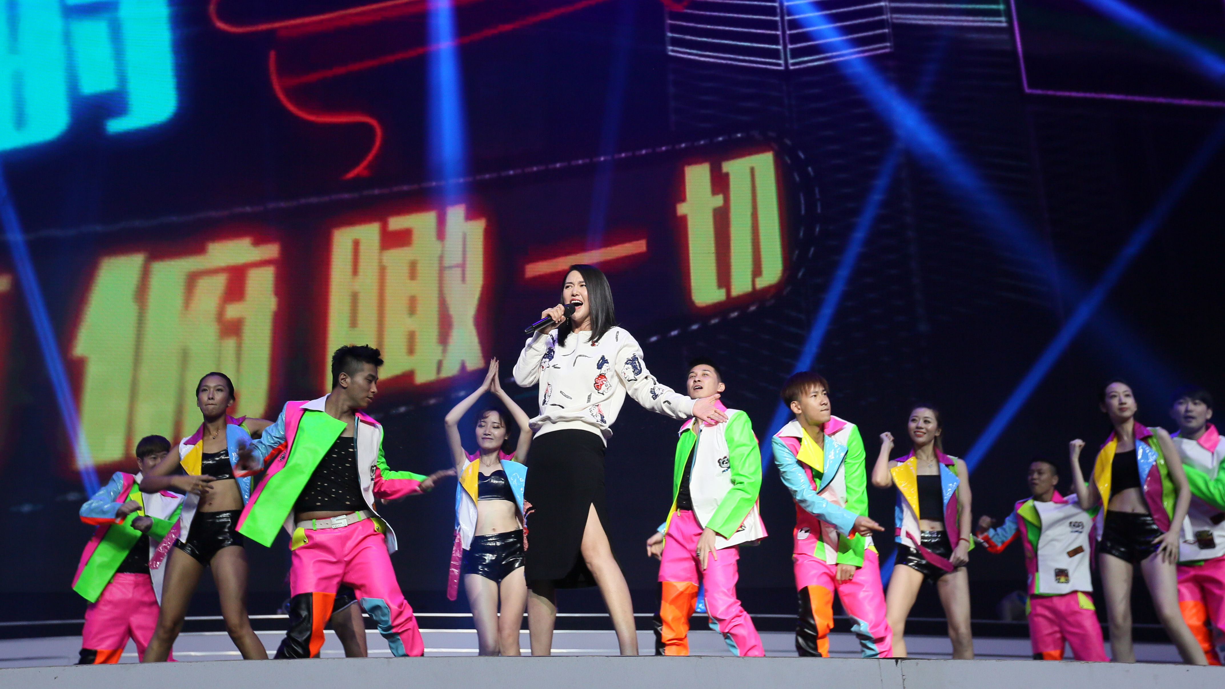 安利活动现场，女歌手立于舞台中央持麦倾情演唱，多名身着炫彩潮流服饰的舞者同步伴舞，背景大屏显示“俯瞰一切”字样，舞台灯光酷炫灵动，现场氛围热烈昂扬，尽显活动的活力与感召力。