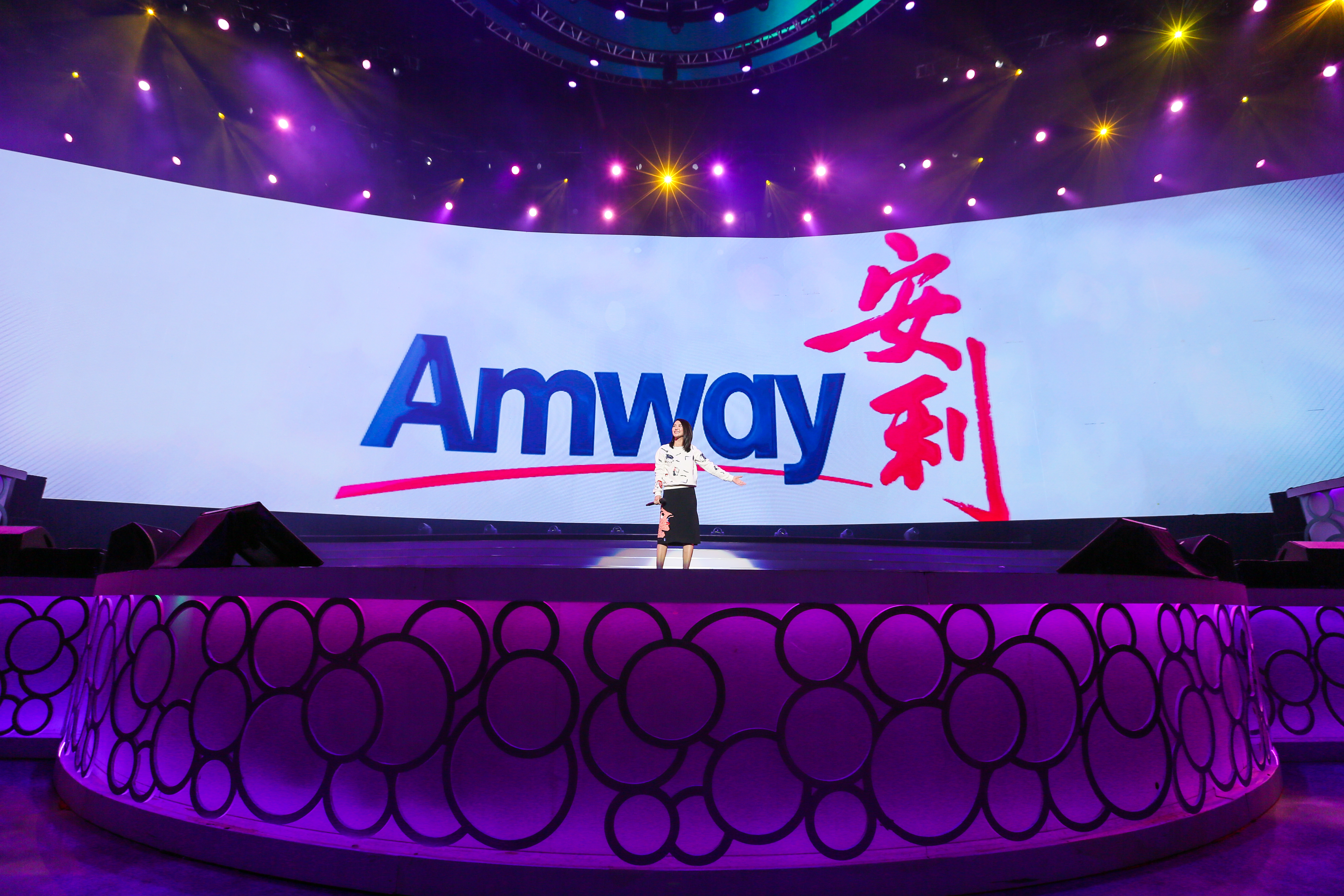 活动现场灯光绚烂，弧形巨屏上清晰呈现“Amway”“安利”品牌标识，一位女性嘉宾站在设计精致的舞台中央，手持话筒舒展手臂进行分享，现场氛围热烈积极，充分展现安利品牌活动的专业活力与昂扬风貌。