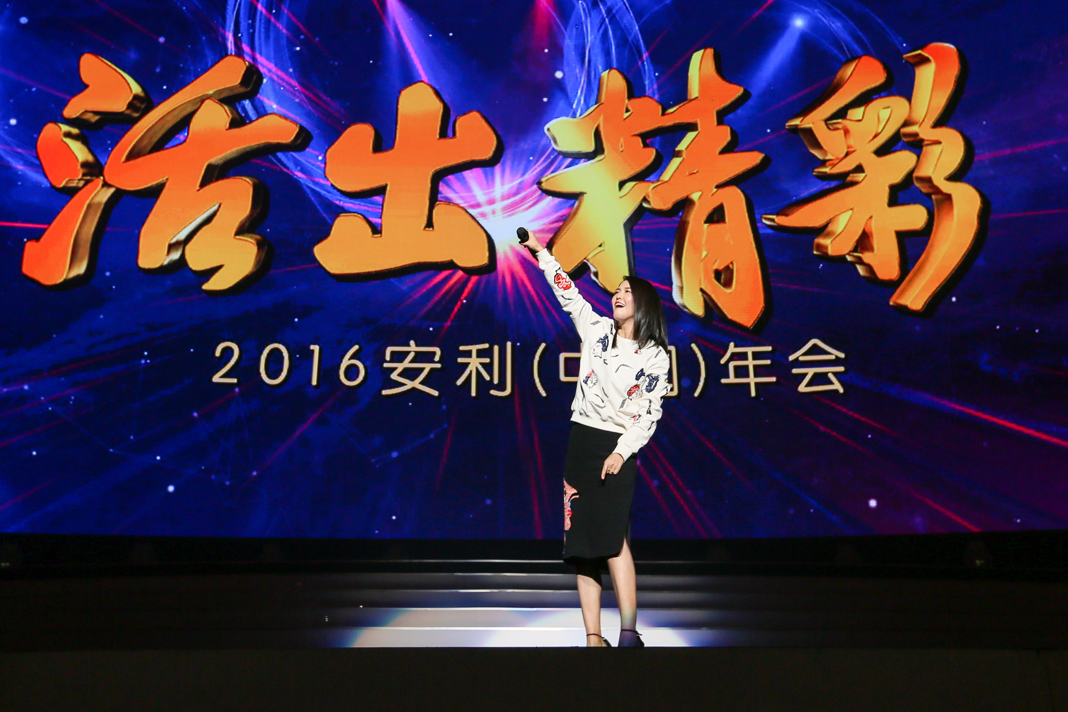 本次为2016安利（中国）年会活动现场，背景大屏醒目展示“活出精彩”主题字样，光影效果绚丽。女嘉宾站在舞台上抬手与台下互动，神情昂扬愉悦，现场氛围热烈积极，充分传递出安利品牌的正向活力。
