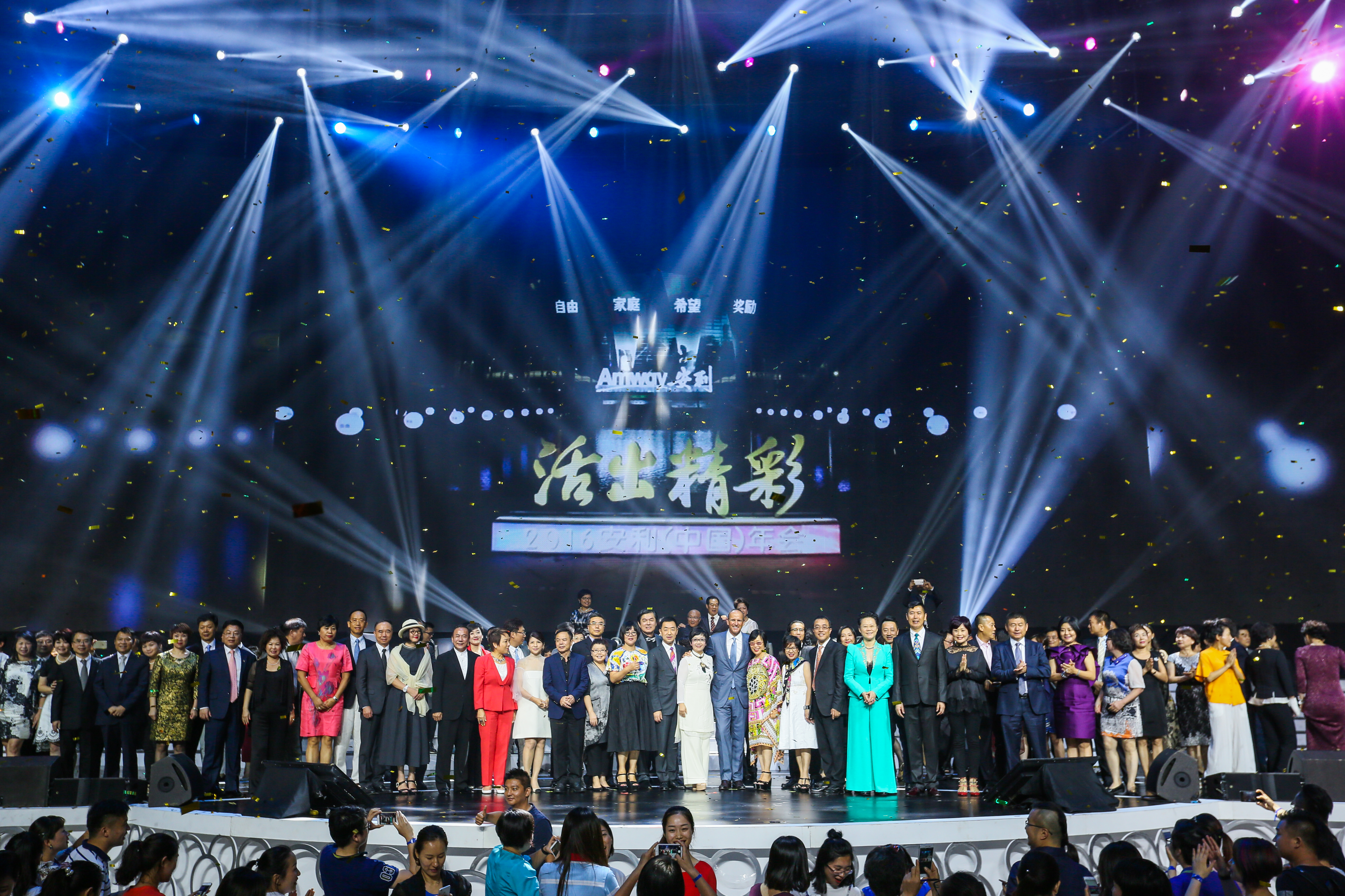 这是2016安利（中国）年会现场，背景大屏展示安利核心价值观“自由、家庭、希望、奖励”、品牌标识及“活出精彩”主题字样。舞台灯光绚烂，金色彩屑飘落，众多伙伴在舞台整齐列队合影，台下观众纷纷记录，现场氛围热烈昂扬，尽显品牌正向活力。