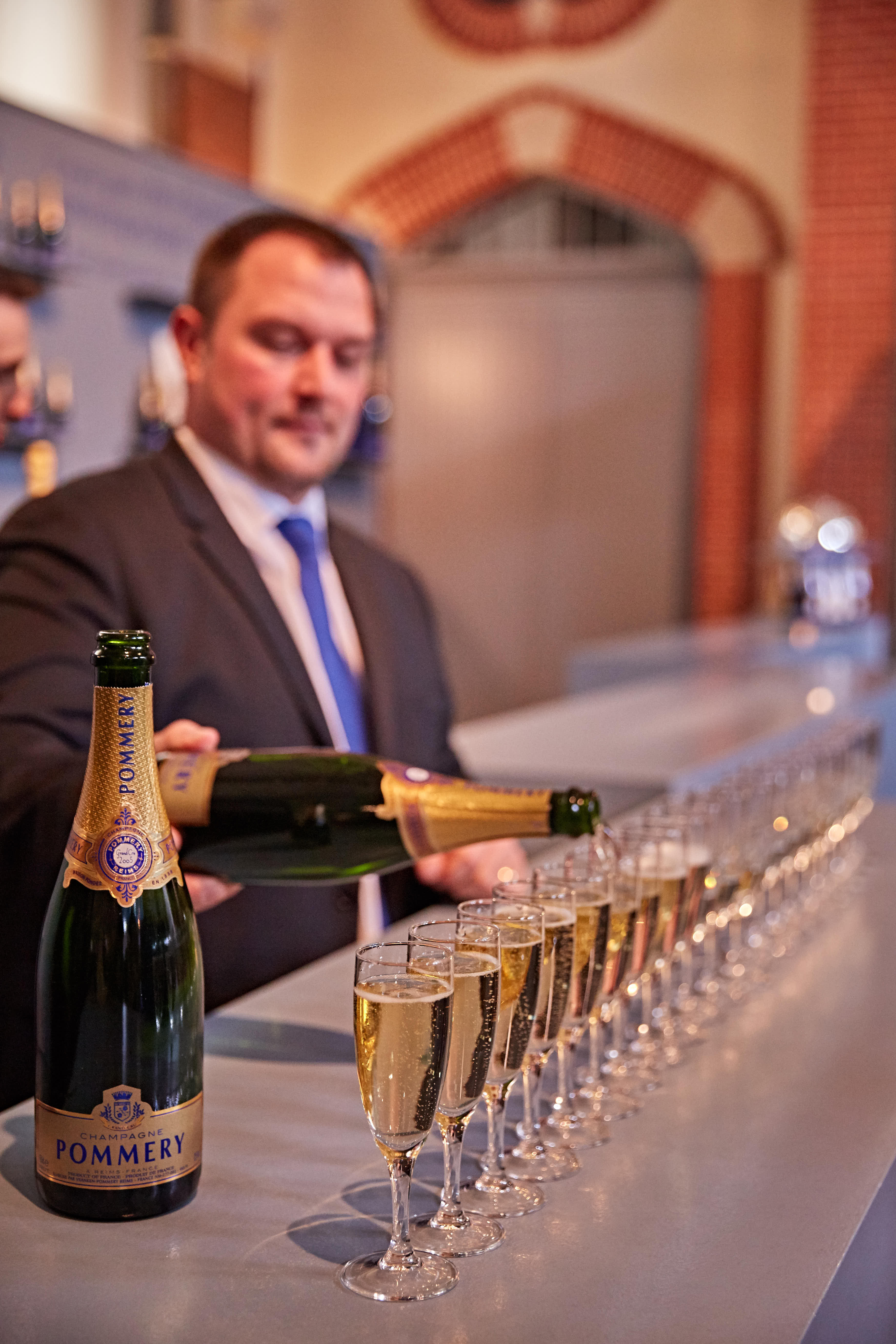 画面中工作人员正将POMMERY香槟依次斟倒入吧台上整齐排列的高脚杯中，金黄酒液泛着细密气泡，场景精致考究，烘托出安利品牌活动正式愉悦的氛围，为后续庆祝环节做好充分准备。
