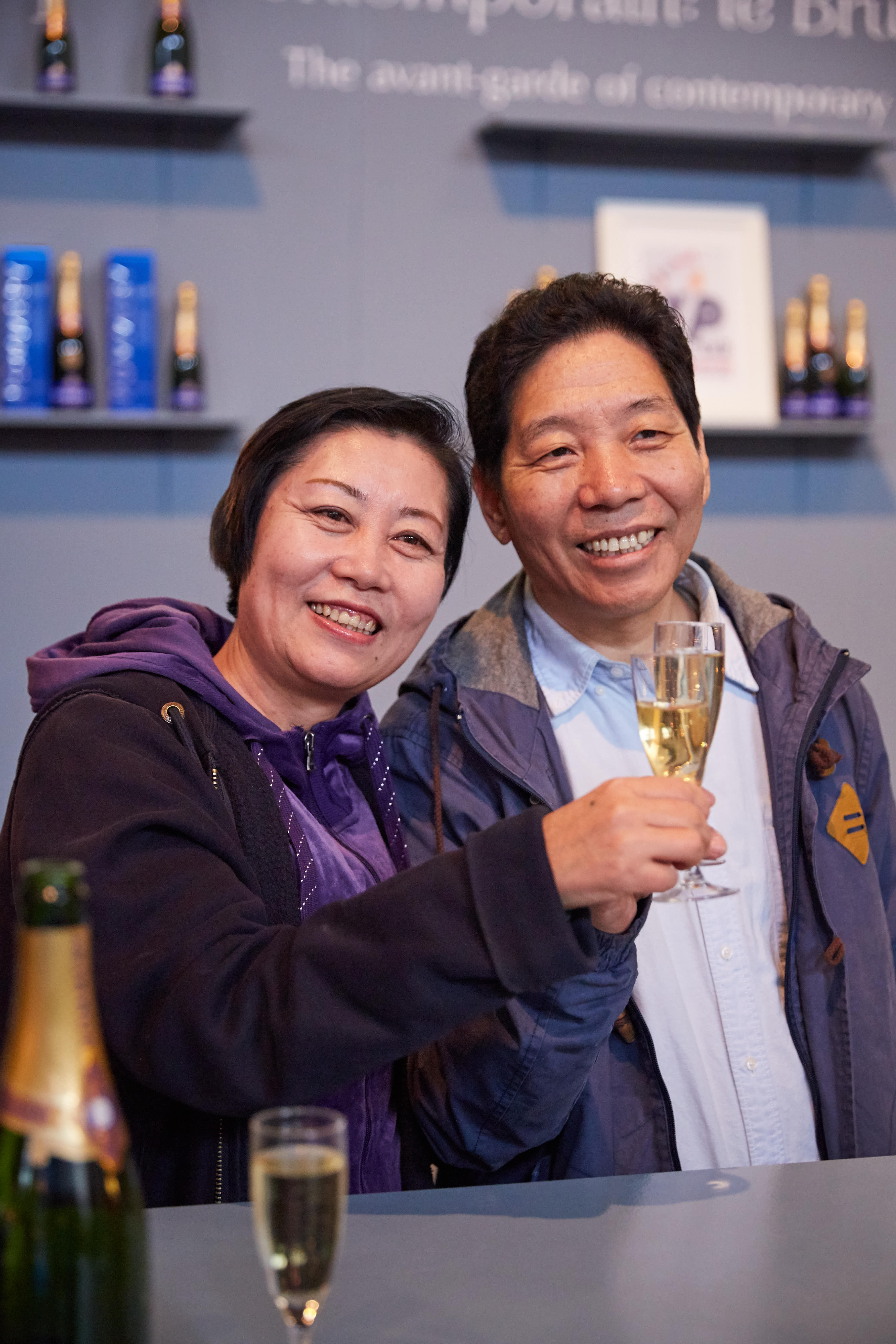 活动现场，两位安利伙伴笑容和煦，共同举起盛有香槟的高脚杯，背景置物架上陈列着酒品与相关陈设，整体氛围轻松愉悦，尽显伙伴们共贺收获的满满喜悦，烘托出活动温馨融洽的欢庆氛围。