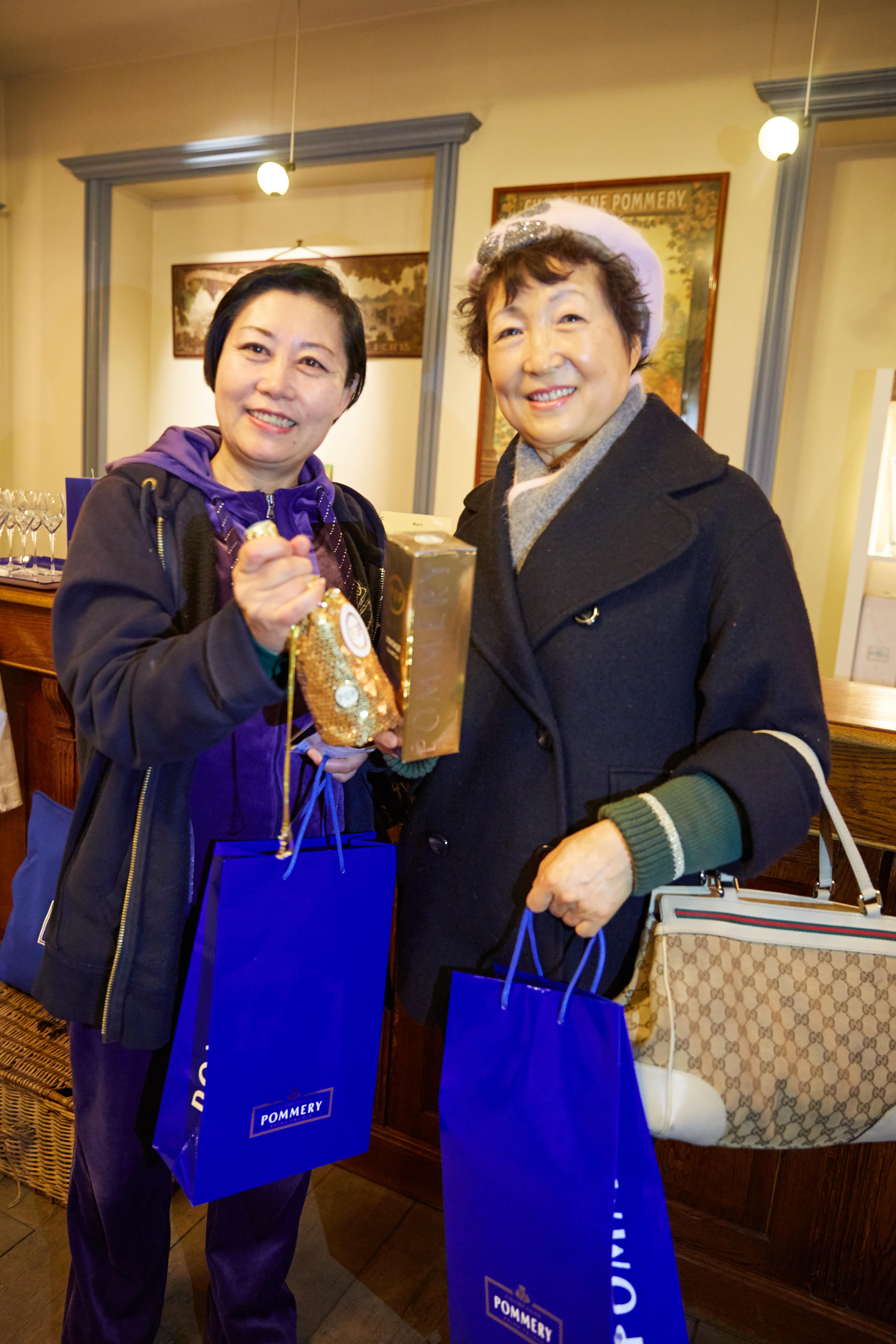 安利线下活动现场暖意融融，两位笑容灿烂的伙伴手持印有“POMMERY”标识的蓝色定制礼品袋，其中一位伙伴正展示获赠的精美礼品，氛围轻松愉悦，尽显活动的贴心福利与伙伴们的喜悦心情，传递出满满的幸福感。