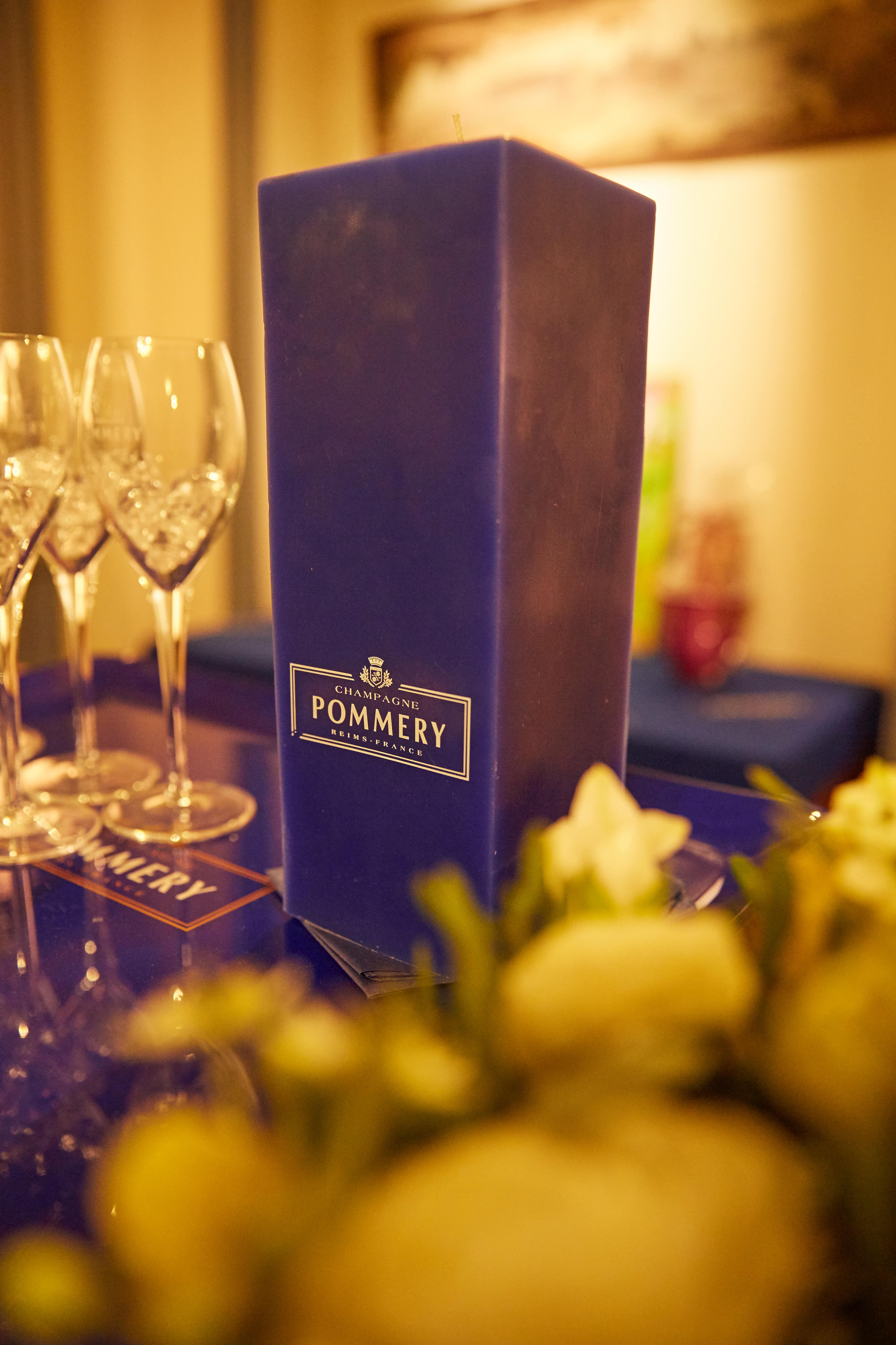 暖调光影下，活动酒会区域桌面摆放着印有“CHAMPAGNE POMMERY REIMS-FRANCE”标识的香槟礼盒，旁侧整齐陈列着剔透的香槟杯，前景以清新花艺点缀，整体氛围雅致高端，尽显活动精致质感。