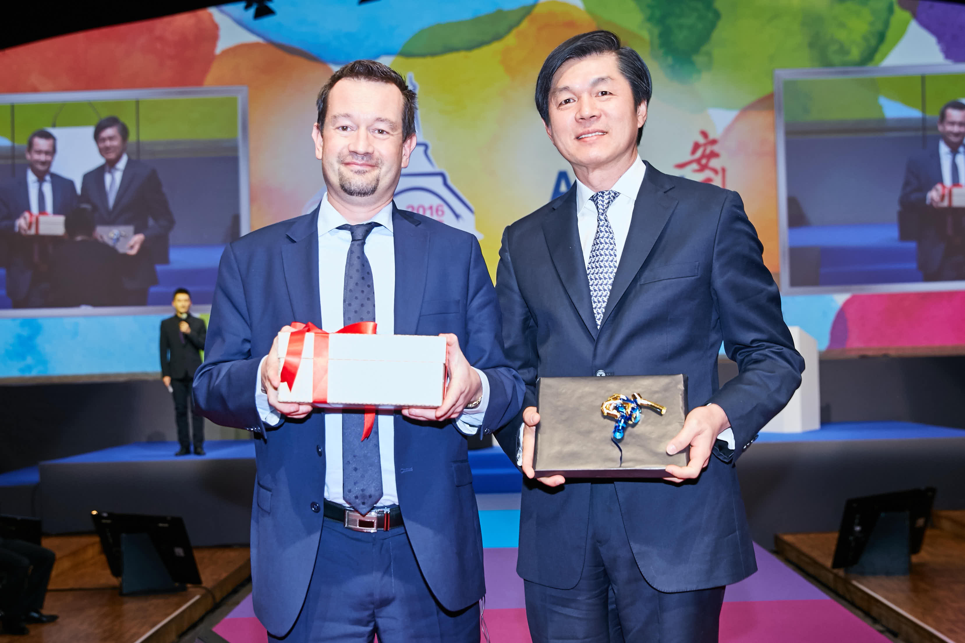 这是2016年安利品牌活动现场，两位身着正装的活动代表站在舞台上，分别手持系有红丝带的礼盒、带有精美装饰的纪念礼盒，背景是色彩明快的活动主题大屏，现场氛围正式融洽，充分展现双方友好协作的积极姿态，凸显活动纪念价值。