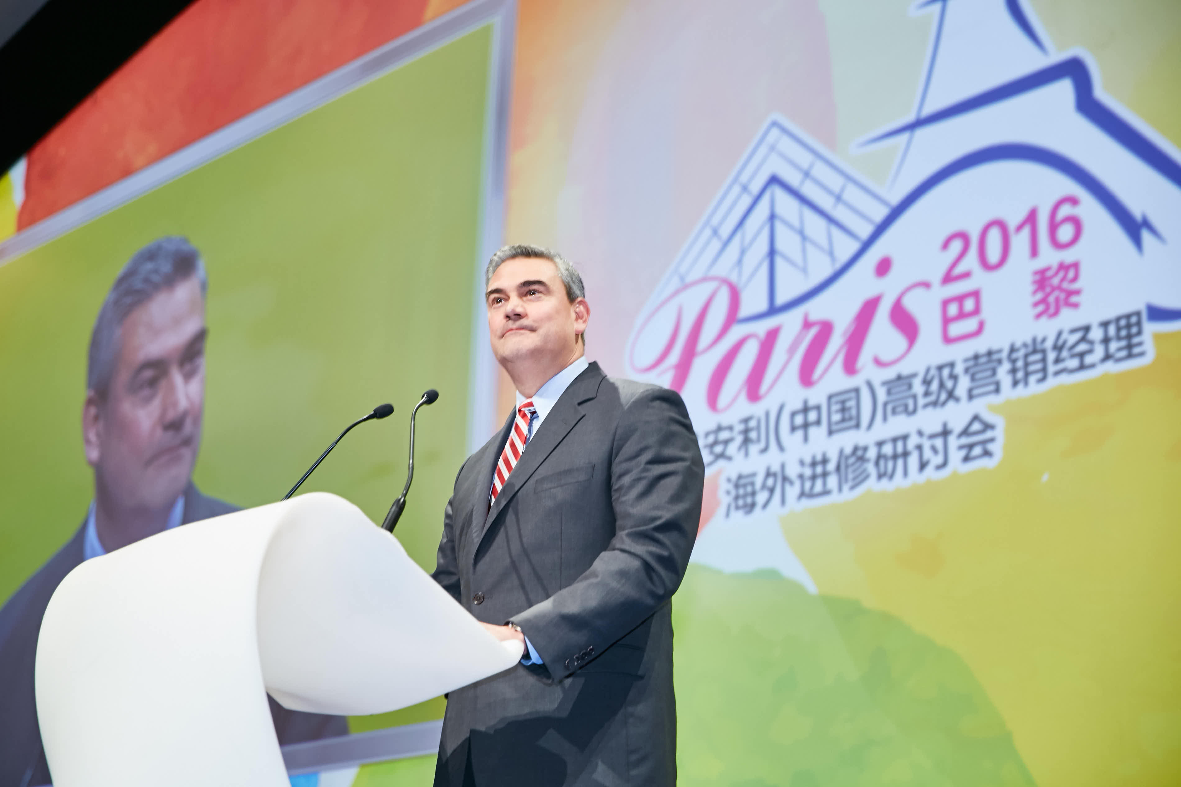 图为2016巴黎安利（中国）高级营销经理海外进修研讨会现场，身着正装的外籍高管站在白色发言台后发表致辞，背景大屏印有活动专属标识与嘉宾形象，整体氛围正式庄重，充分彰显活动的专业属性与高端定位。