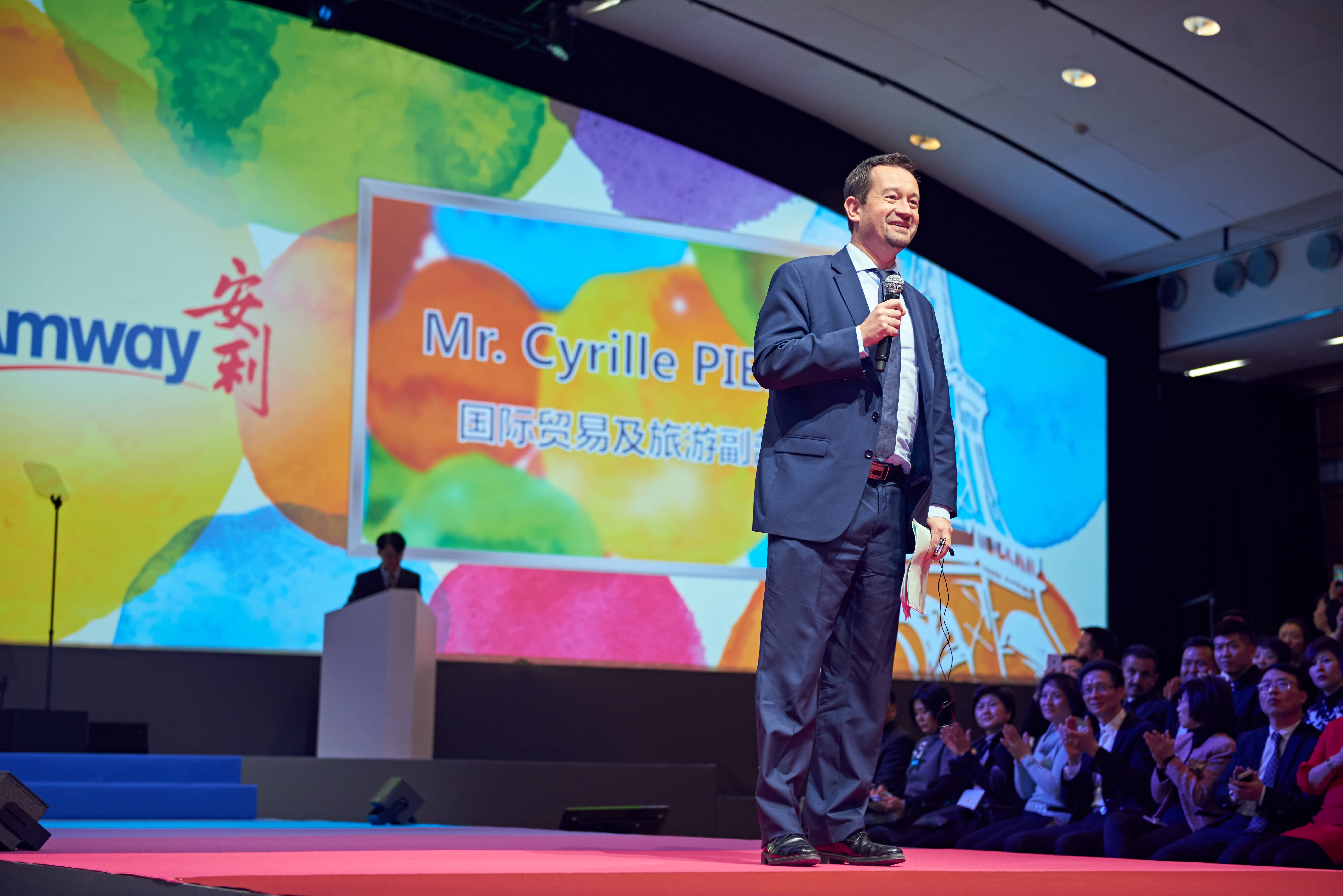 本次安利品牌活动现场，国际贸易及旅游相关官员Mr. Cyrille PIE身着正装持话筒登台致辞，身后大屏清晰展示安利品牌标识与嘉宾身份信息，台下参会伙伴纷纷鼓掌，现场氛围热烈昂扬，彰显活动高规格与安利积极的品牌风貌。