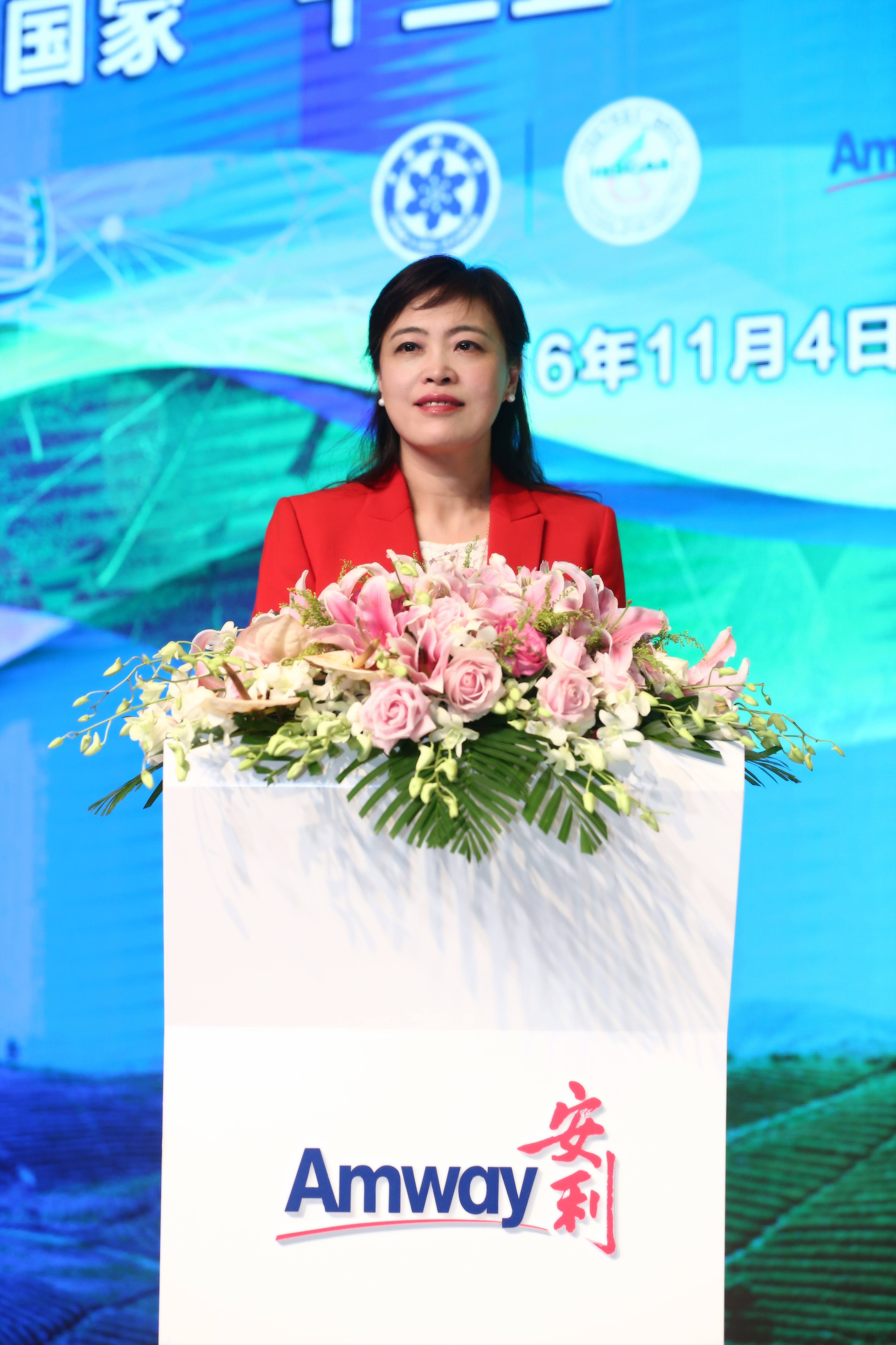 本次活动于2016年11月4日举办，身着红色正装的女士站在印有“Amway 安利”标识、装饰有粉百合、玫瑰等精美花艺的讲台后发言，背景大屏展示相关合作标识与活动视觉元素，现场氛围庄重专业，充分展现安利品牌活动的正式风貌。