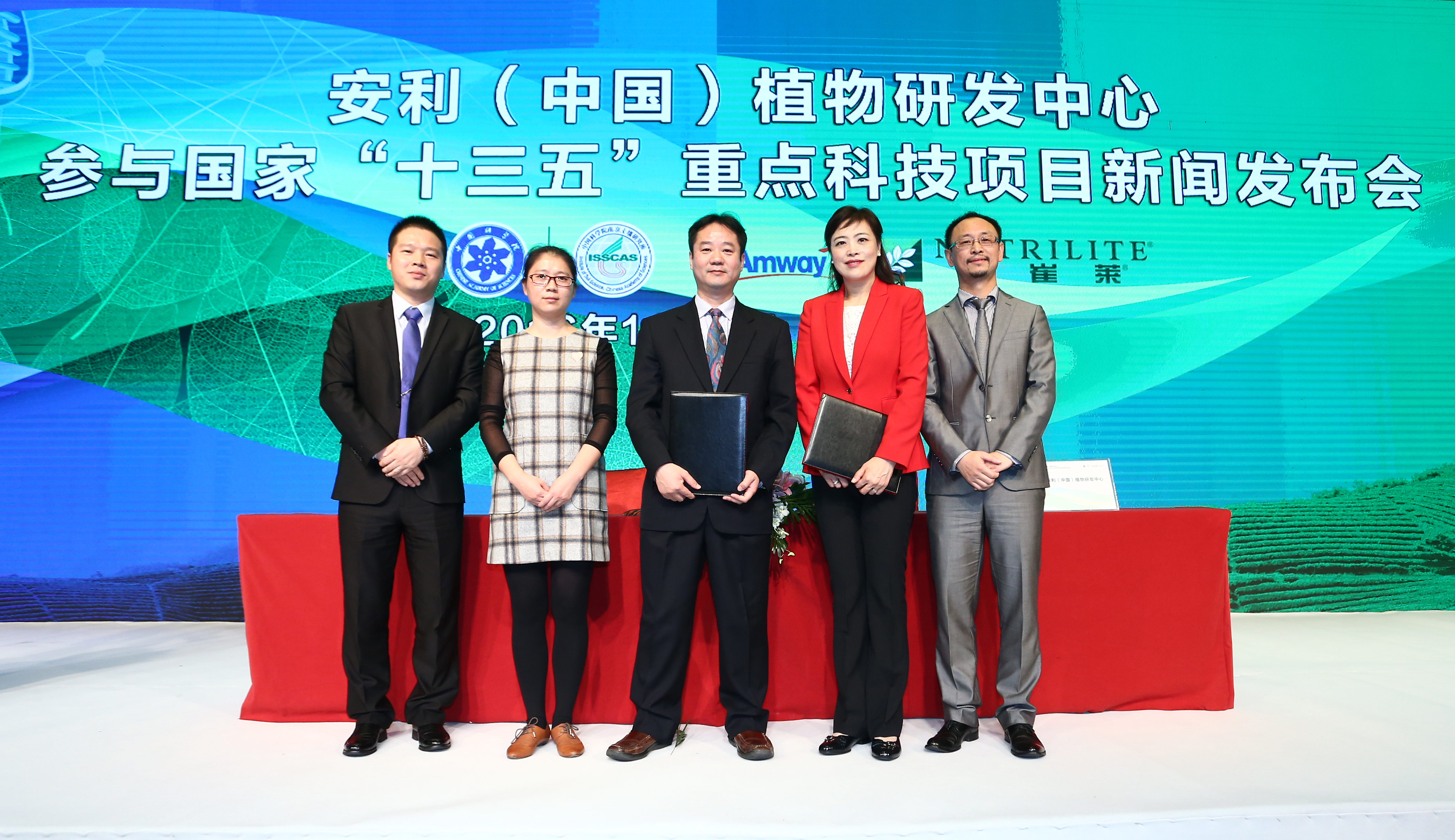 本次为安利（中国）植物研发中心参与国家“十三五”重点科技项目新闻发布会现场，五位嘉宾在签约台后合影，两位项目代表手持签约文本，背景可见安利、纽崔莱及合作科研机构标识，现场氛围庄重正式，彰显安利在植物研发领域的专业实力。