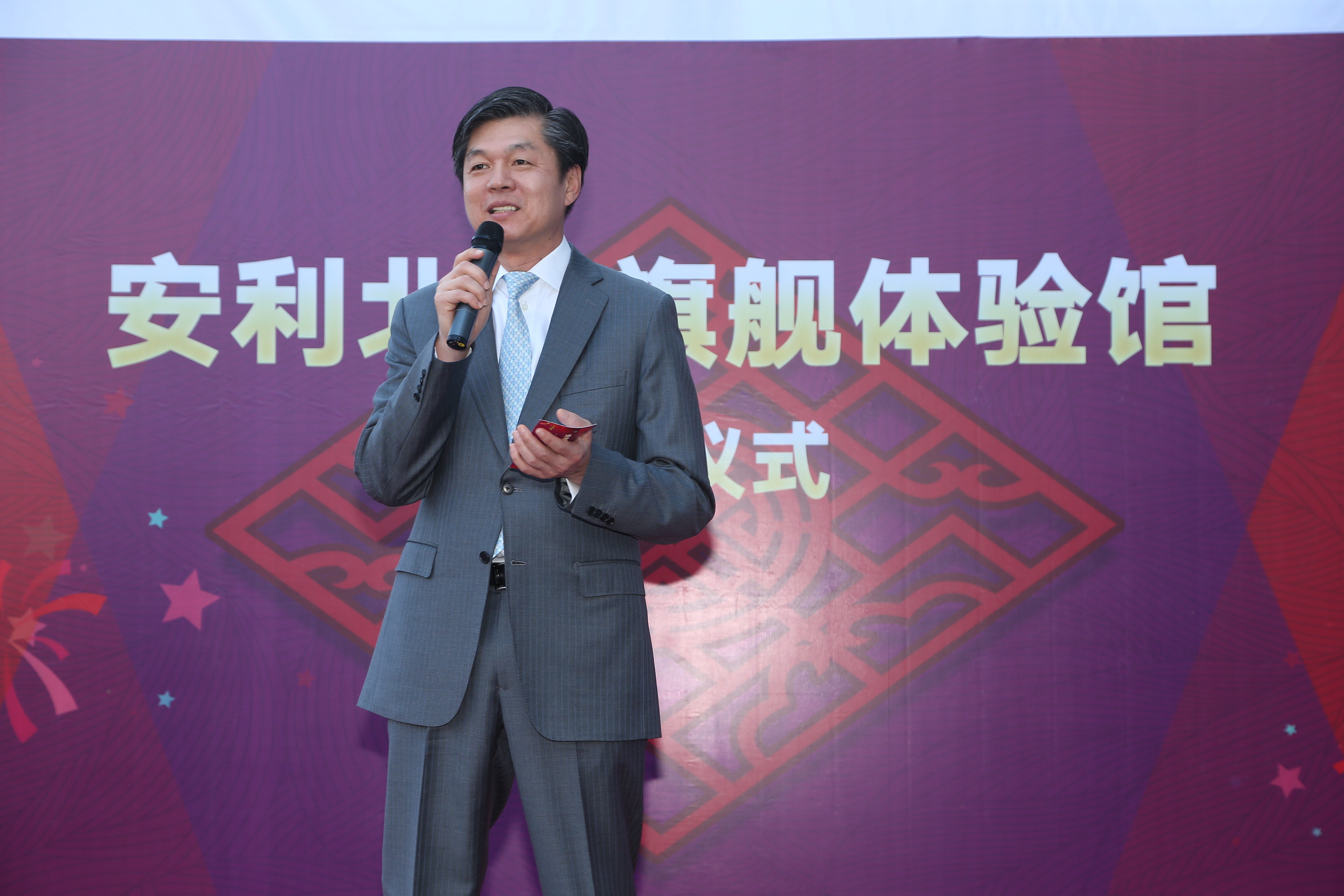 画面中身着灰色正装的男士手持话筒与红色手卡正在发言，背景为印有“安利北京旗舰体验馆”“仪式”字样的活动主题背景板，现场氛围正式隆重，充分展现安利品牌活动的专业风貌。