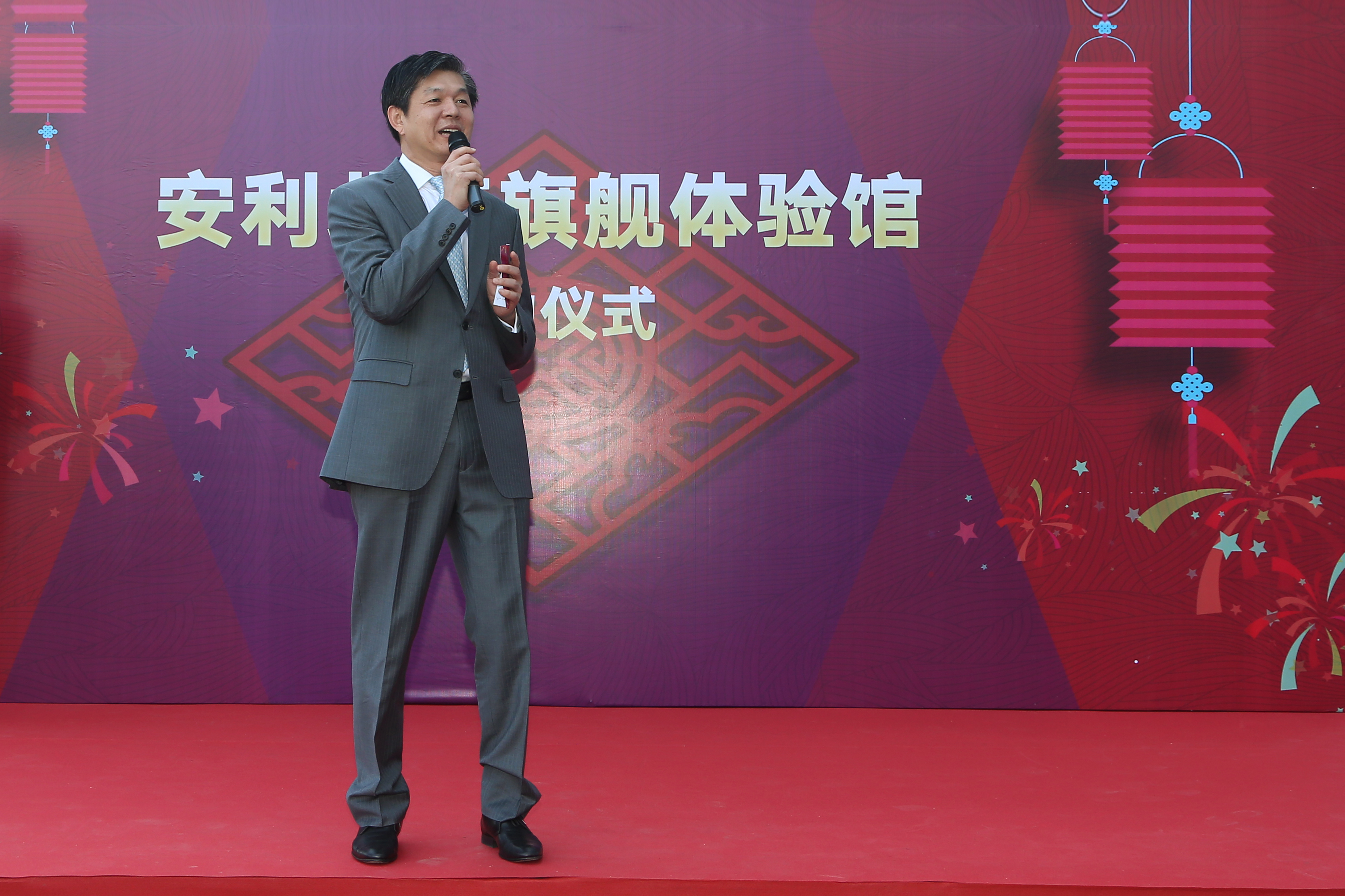 本次是安利旗舰体验馆相关仪式现场，身着灰色正装的嘉宾手持话筒站在红色舞台上致辞，背景板以红紫为主色调，搭配灯笼、烟花等喜庆装饰，印有“安利旗舰体验馆”“仪式”字样，现场氛围庄重喜庆，尽显安利品牌活动的积极风貌。