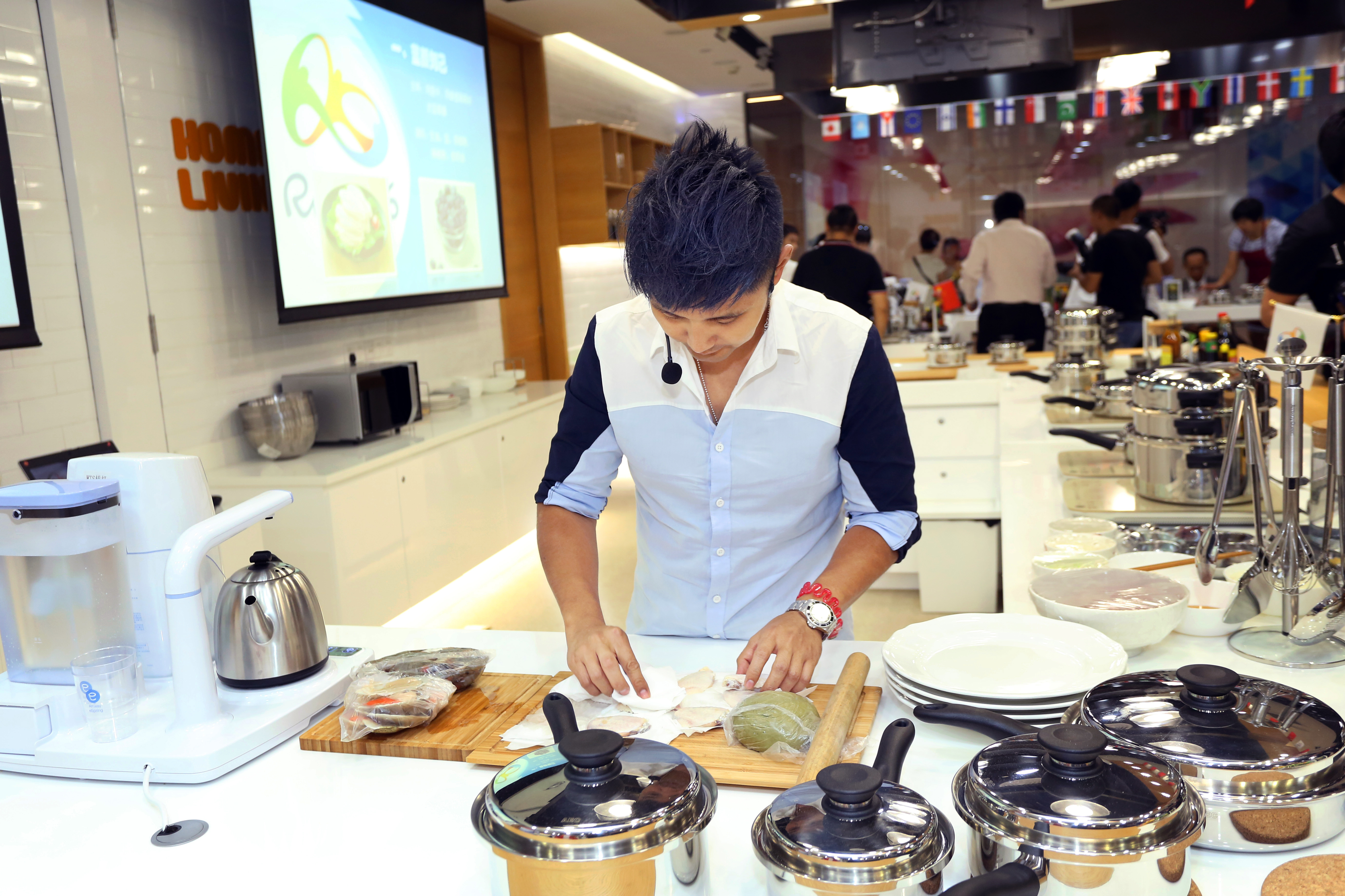 本次是安利HOME LIVING主题美食体验活动，专业讲师正于操作台前专注处理食材，台面摆放着安利皇后锅具、净水设备等家居科技产品，背景显示屏展示食谱内容，现场氛围活跃，充分传递安利健康生活的品牌理念。