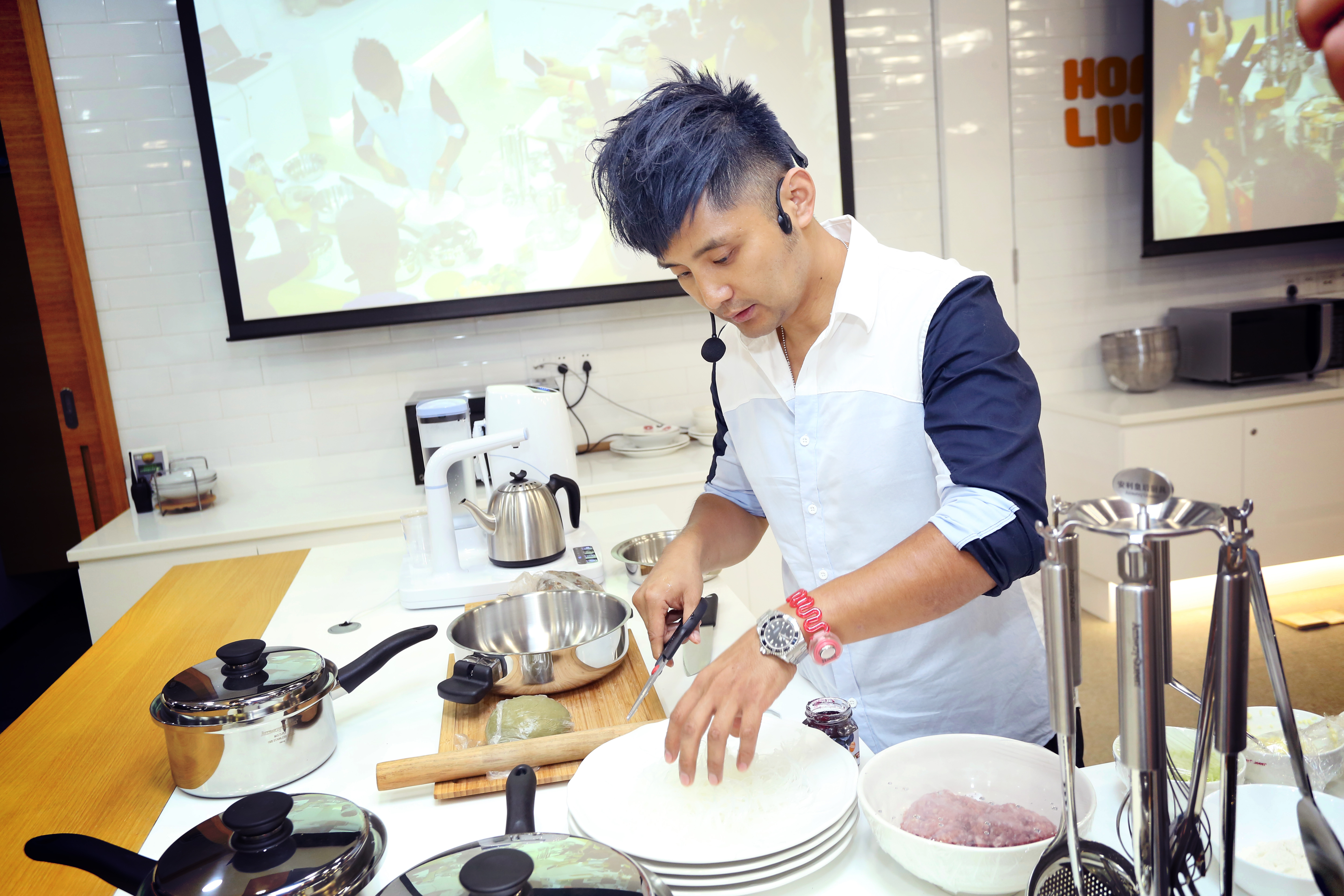 佩戴耳麦的专业讲师在整洁的料理操作台前演示美食制作流程，台面上摆放着安利皇后系列锅具、新鲜食材与各类料理工具，背景显示屏同步转播操作细节，现场氛围轻松专业，传递安利倡导健康饮食、美好生活的品牌理念。
